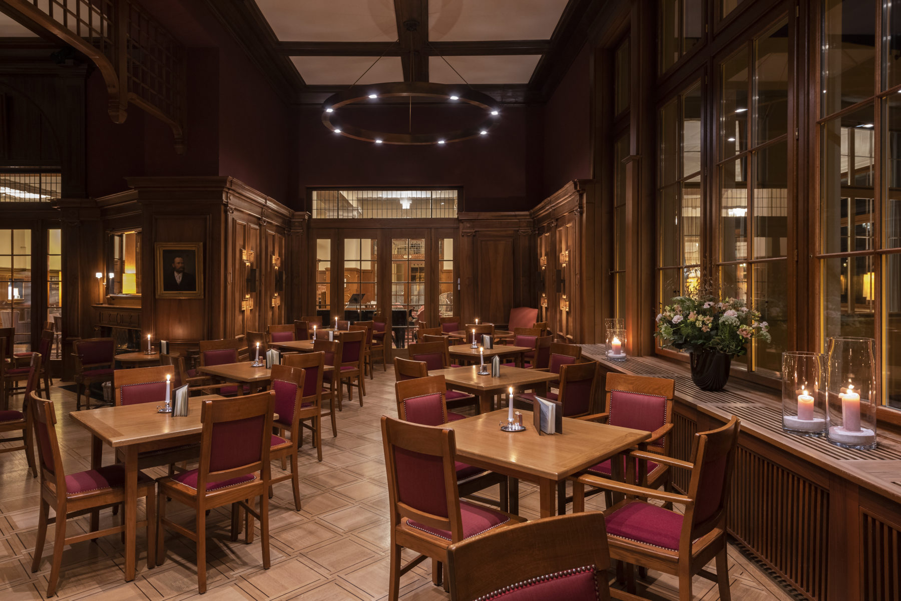 A warm, atmospheric dining room with rich wood wall paneling, parquet flooring, and dark walls. Wooden square tables with lit candles are set with wooden chairs featuring reddish-maroon upholstered seats. Large glass windows line the side, reflecting ambi