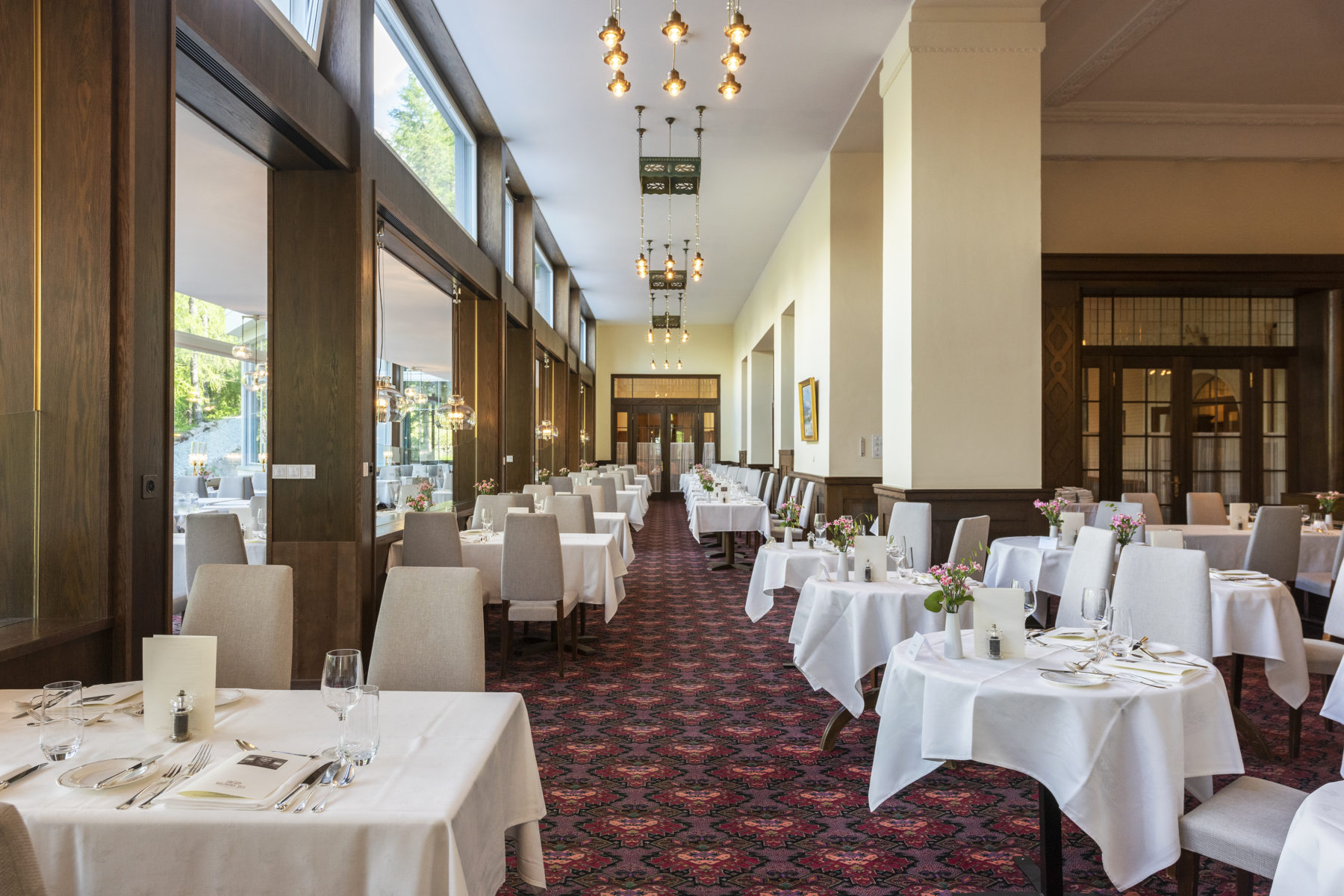 A brightly lit, spacious hotel restaurant featuring a long hallway carpeted in a red patterned rug, lined with dining tables draped in white tablecloths and paired with light grey upholstered chairs. Large windows on the left allow abundant natural light,