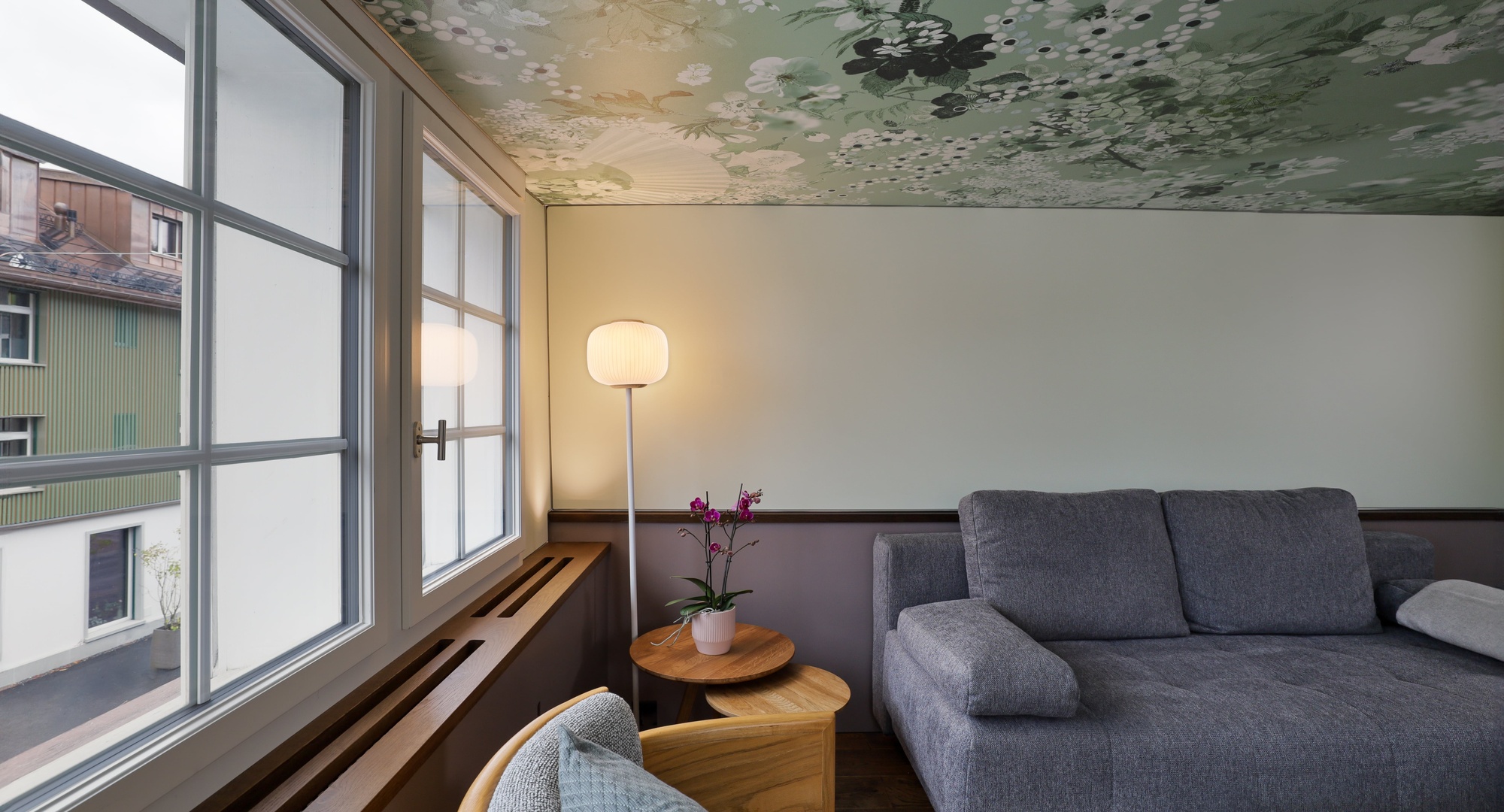 A modern, serene living space featuring a gray fabric sofa, a floral sage green patterned ceiling, large white-framed windows overlooking street architecture, and a glowing floor lamp next to a potted orchid.