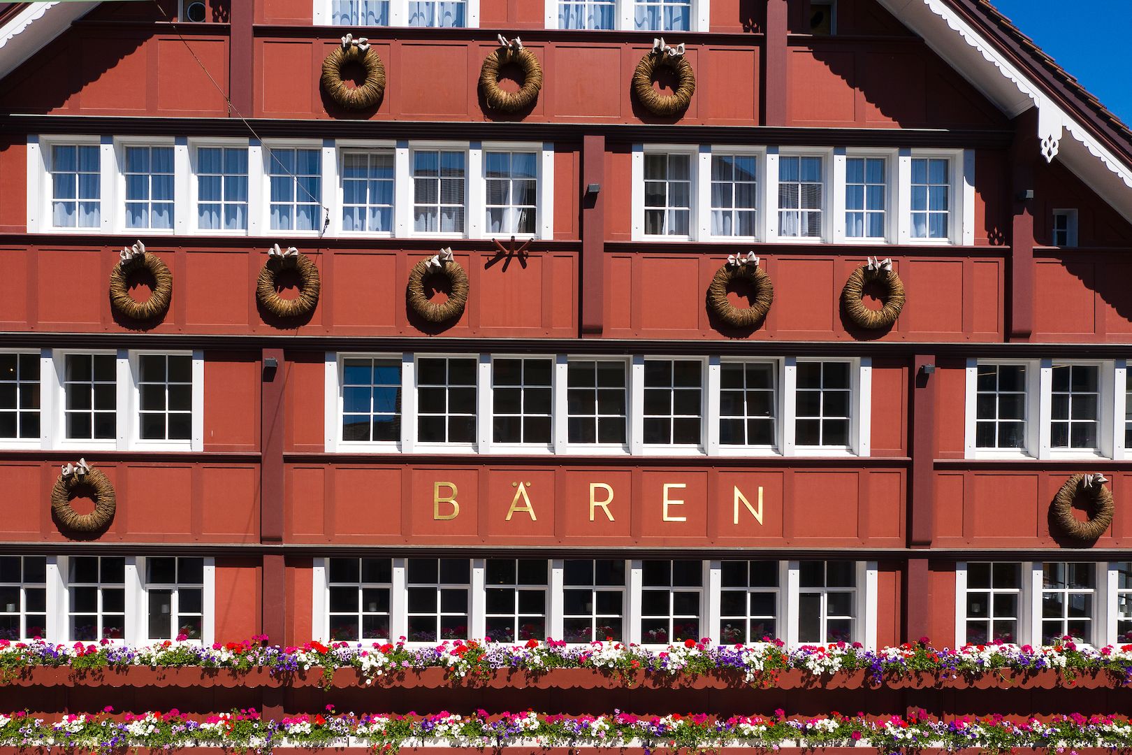 A traditional Swiss alpine building facade painted in rich red, featuring decorative woven wreaths hung between rows of window panes, flower boxes filled with colorful blossoms, and the name 'BÄREN' in gold lettering.
