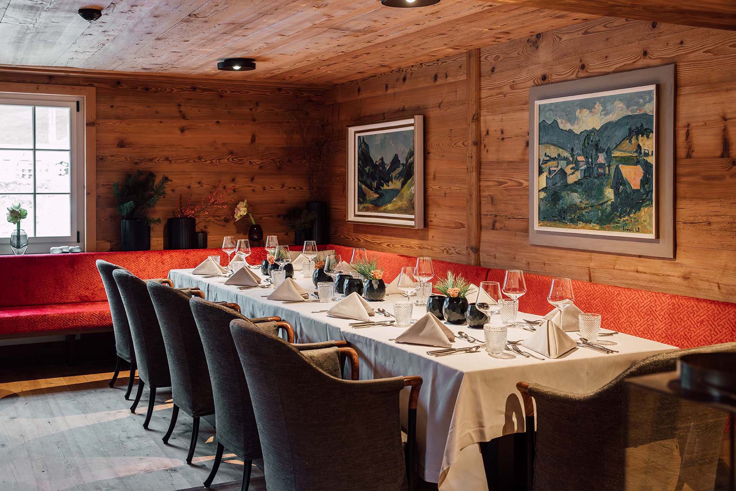 A warm, cozy private dining room clad in wood paneling, featuring a long table dressed in a white tablecloth, grey upholstered armchairs, a bright red corner banquette, fine wine glasses, and framed landscape paintings.
