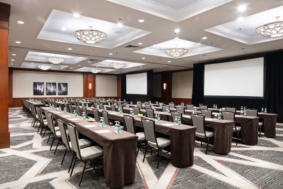 A large conference room set up in a classroom-style layout with rows of long, brown-clothed tables, grey padded chairs, water bottles, and two projection screens at the front.