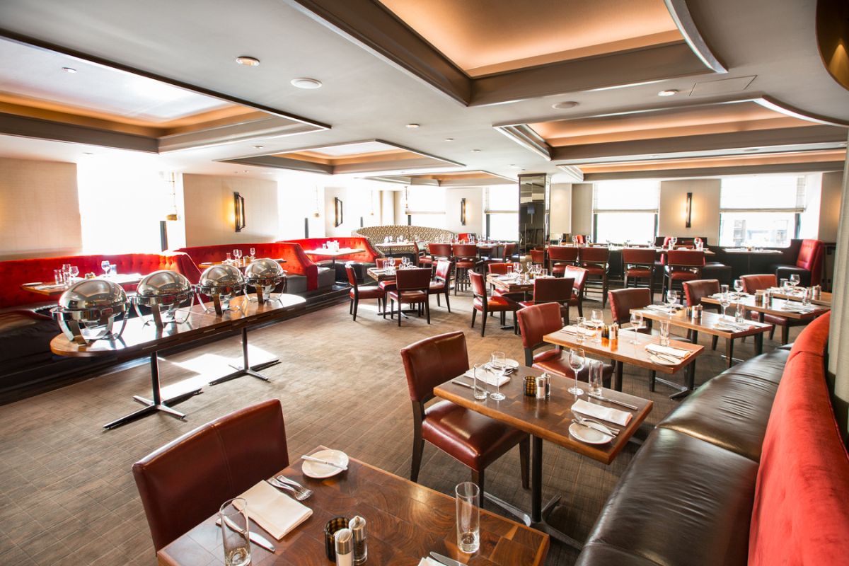 A modern, upscale hotel dining room featuring dark wood tables, reddish-brown leather chairs, long red velvet and leather banquettes, and a buffet station with silver chafing dishes.