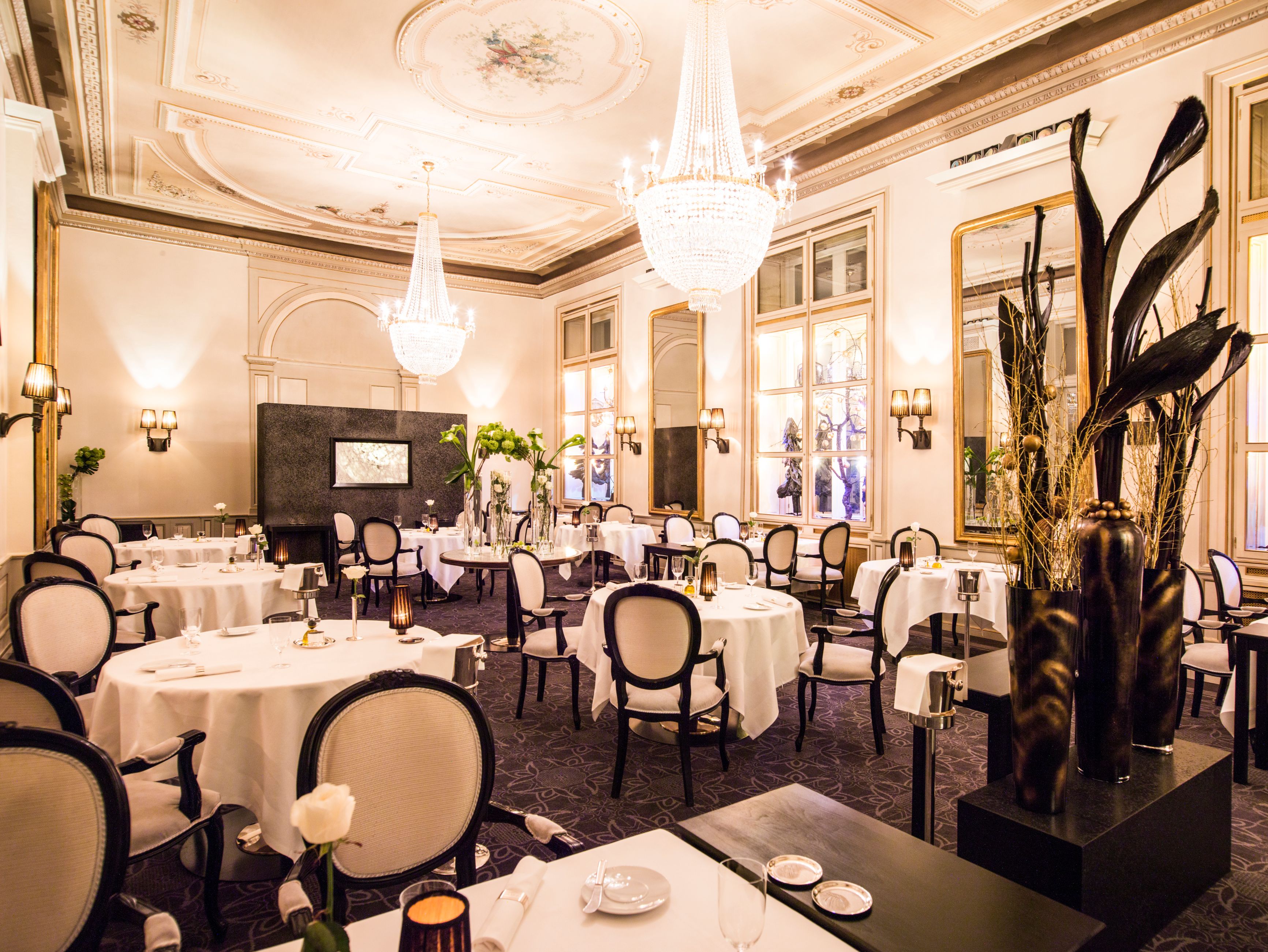A grand, opulent fine dining room with tall ceilings, ornate ceiling frescoes, large crystal chandeliers, round tables with white tablecloths, dark-framed upholstered chairs, and tall decorative floor vases.