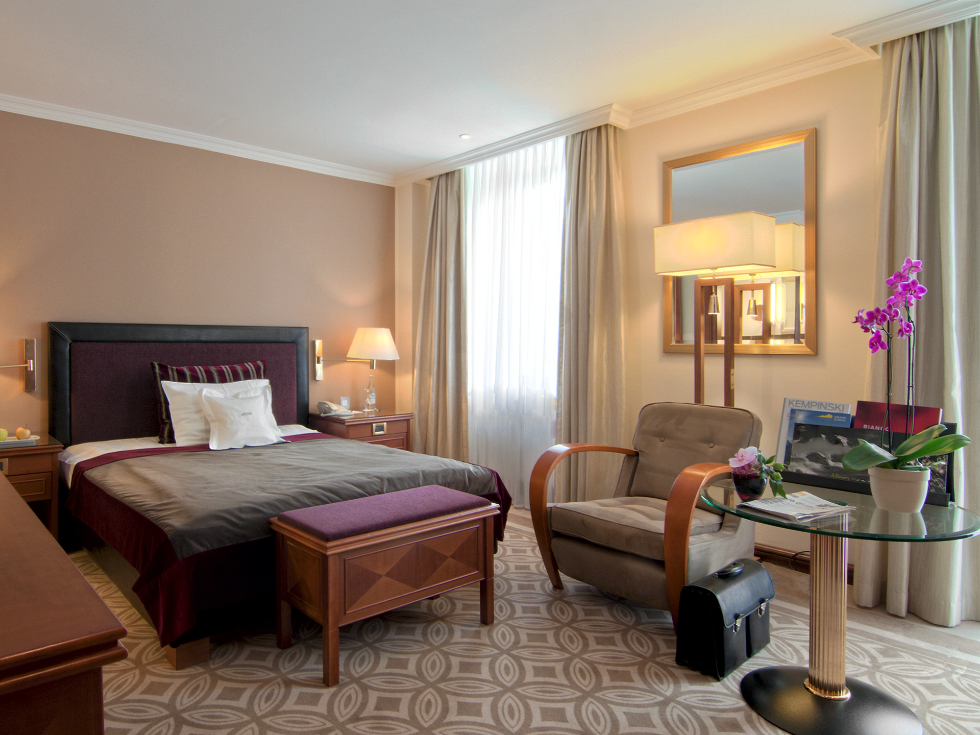 A modern, elegant hotel room featuring a bed with a dark purple headboard and matching bench, a plush lounge armchair with wooden armrests, a round glass side table displaying a potted orchid, patterned beige carpeting, and beige curtains.