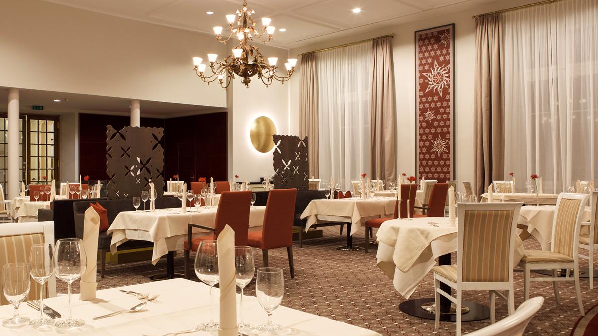 A spacious hotel dining room featuring tables dressed in white linens, warm light brown carpeting, high-backed terracotta red and white striped chairs, sheer beige curtains, and a decorative chandelier.
