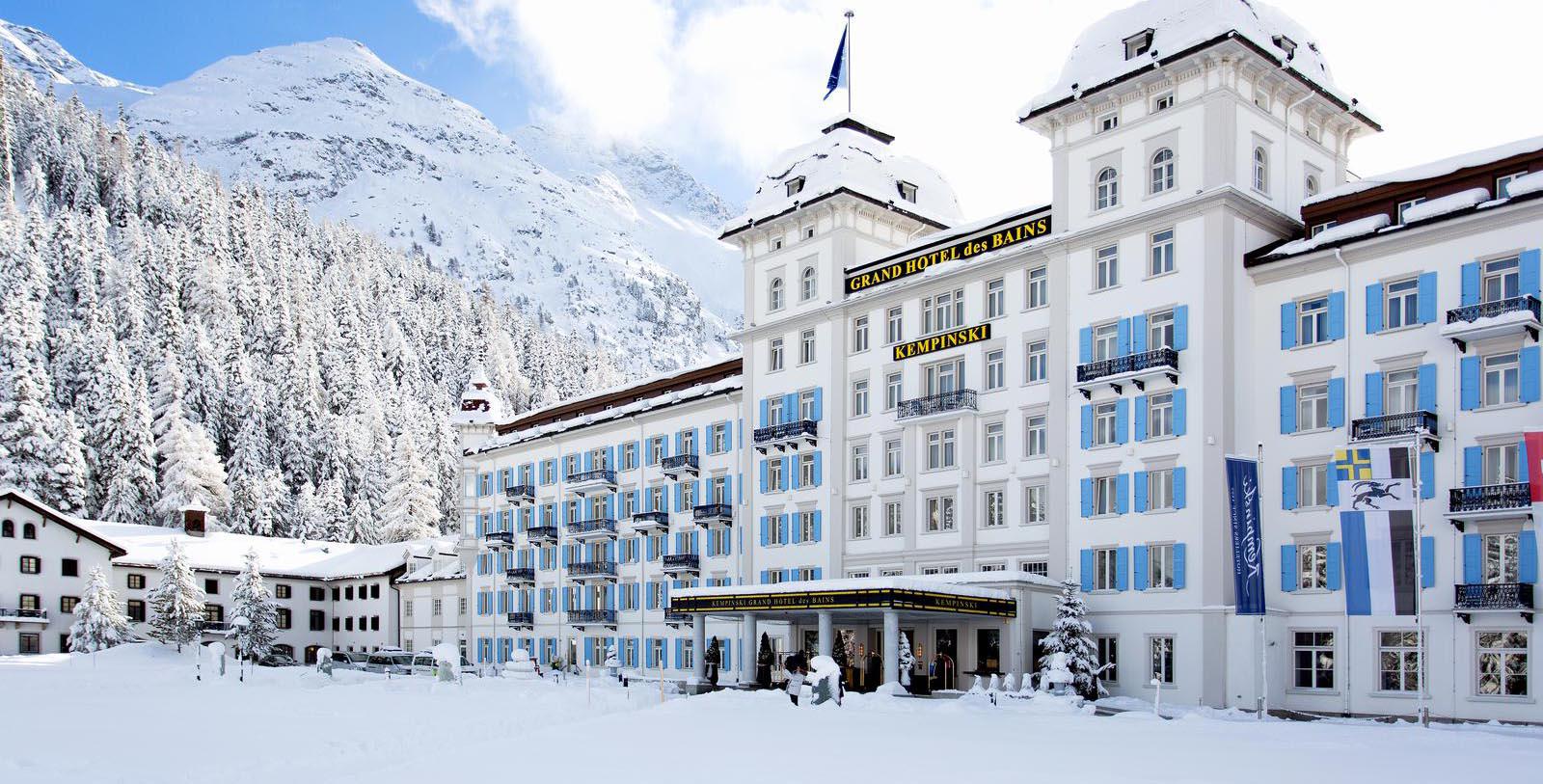 a large white building with blue shutters and a snowy mountain