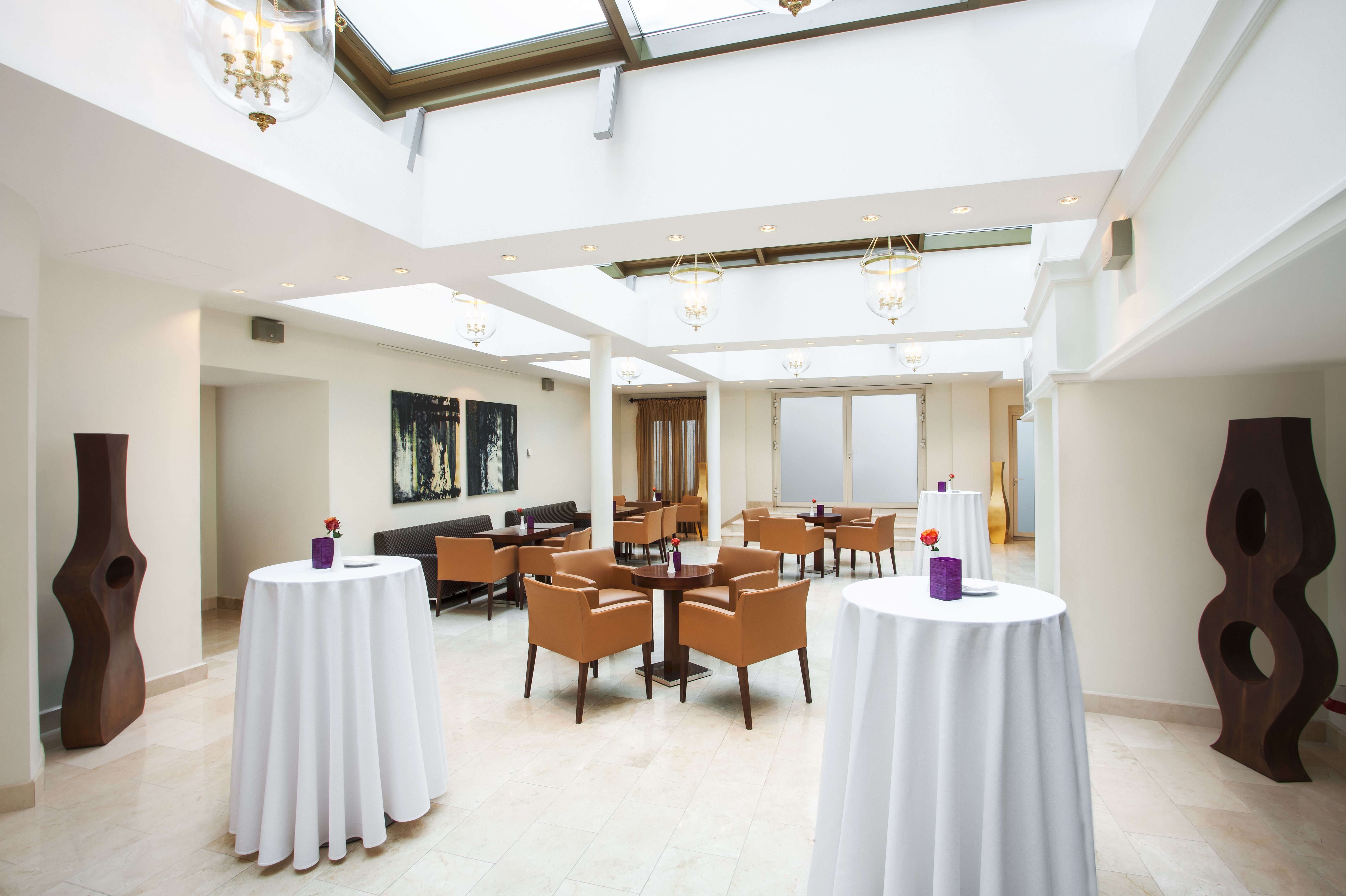 A bright, airy hotel atrium lounge with high glass skylights, high-top cocktail tables wrapped in white linens, tan leather armchairs, and dark sculptural art pieces flanking the room.