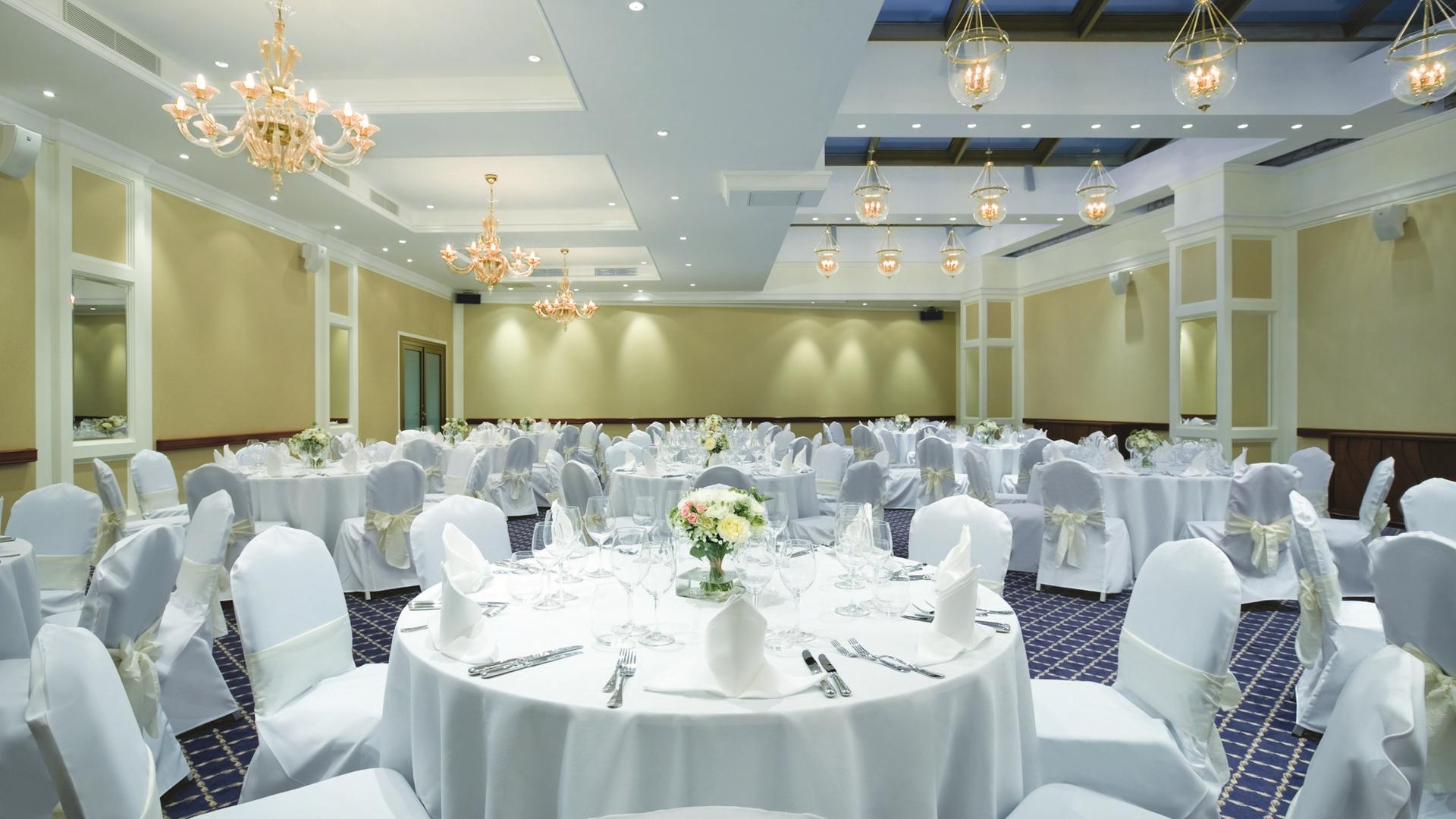 A spacious, elegant banquet hall set up for a formal reception, with round tables adorned with white tablecloths, flower centerpieces, white chair covers, and intricate glass chandeliers illuminated above.