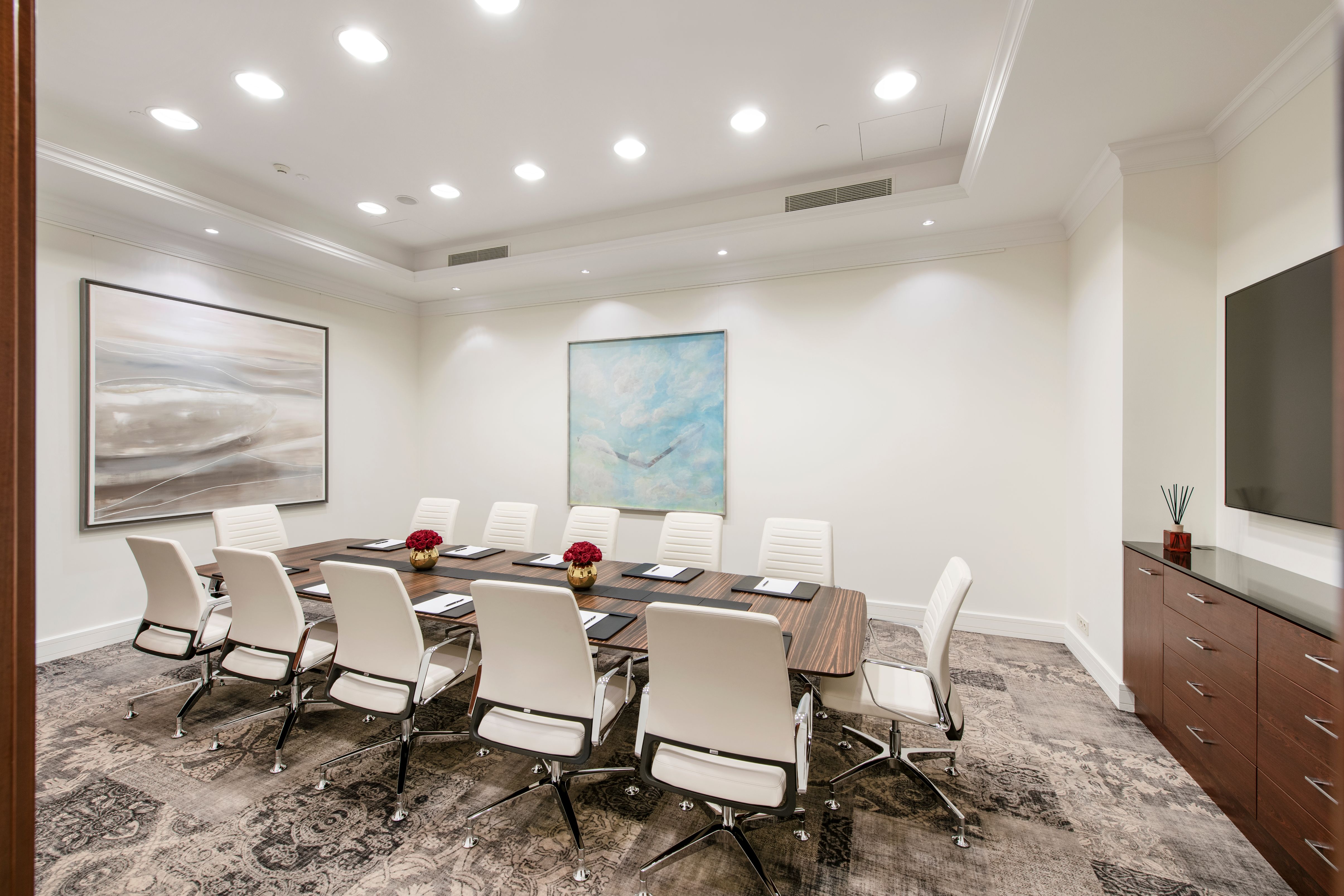 A modern, brightly lit conference room featuring a long wooden board table surrounded by sleek white leather executive chairs, textured patterned carpeting, contemporary paintings, and recessed ceiling lighting.