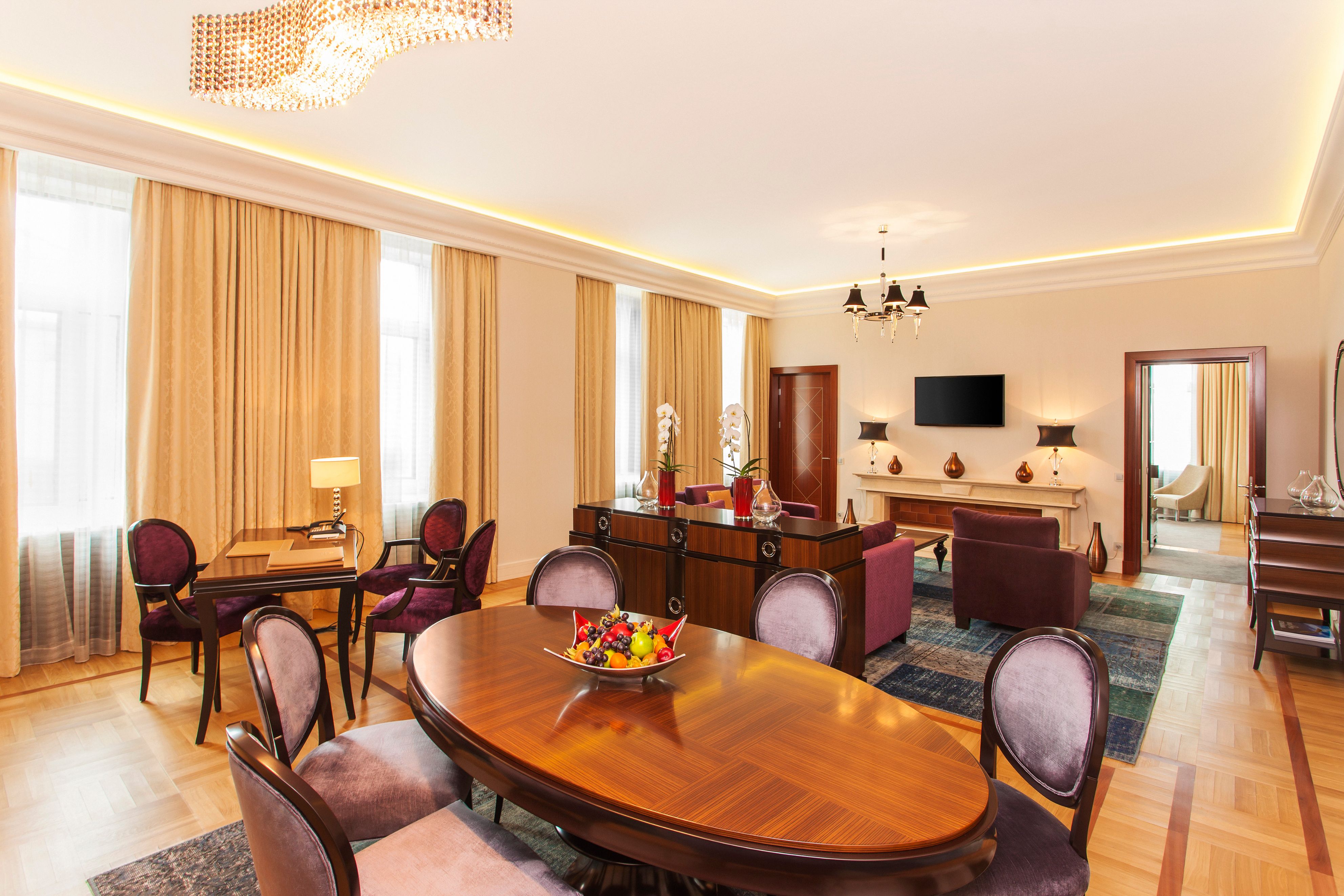 A luxurious hotel suite combining dining and living areas, featuring a large oval wooden dining table with purple velvet chairs, parquet wood flooring, elegant gold drapes, soft cove lighting, and a crystal wave chandelier.
