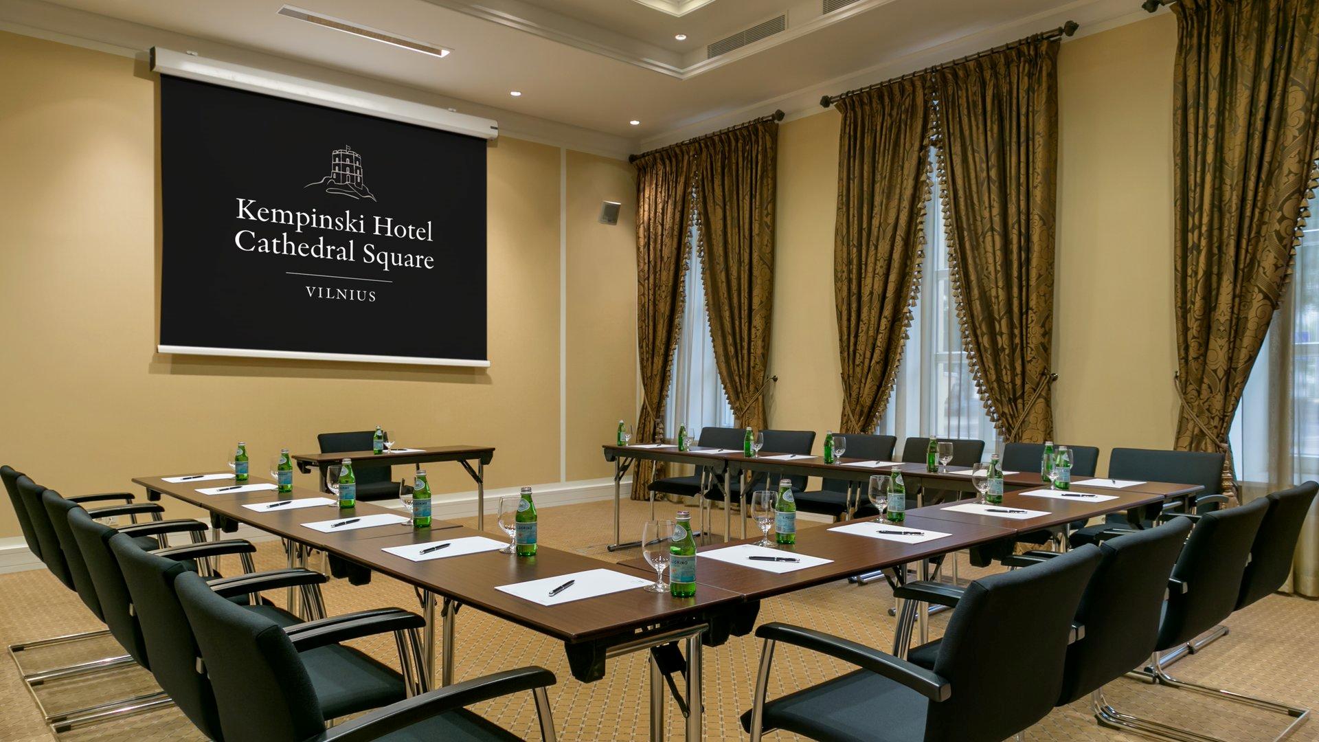 A formal meeting room at Kempinski Hotel Cathedral Square in Vilnius, featuring a U-shape conference table setup with black chairs, bottled water and note pads, heavy patterned gold drapes, and a large projection screen displaying the hotel logo.