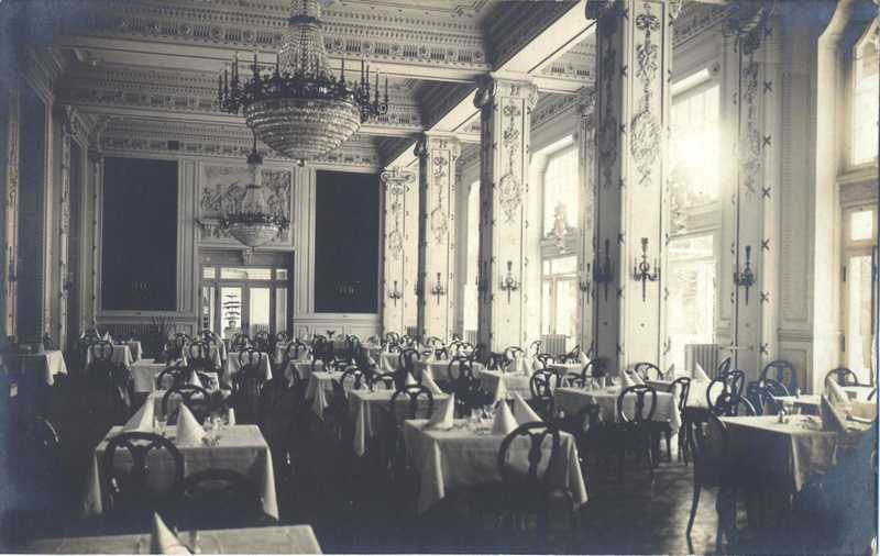 A historic black and white photograph of a grand dining hall featuring high ceilings, ornate pillars, large crystal chandeliers, and multiple rows of set dining tables.