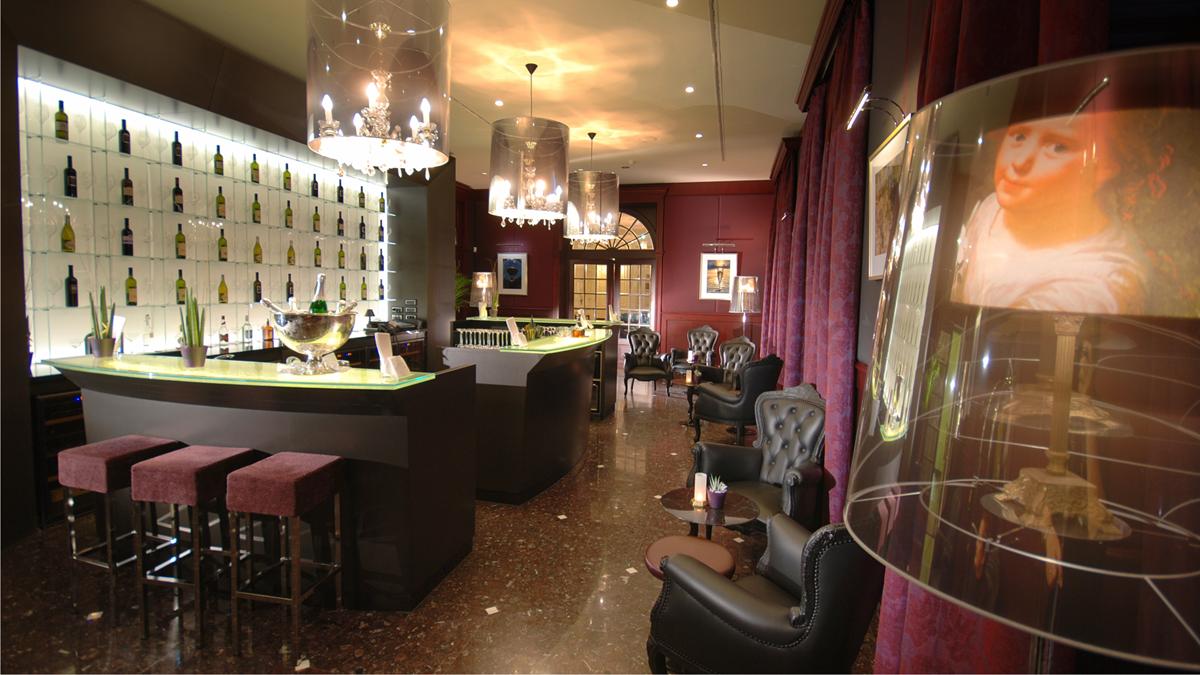 An upscale lounge and bar area with a wall-mounted wine display, modern pendant chandeliers, bar stools, dark leather armchairs, and rich red drapery.