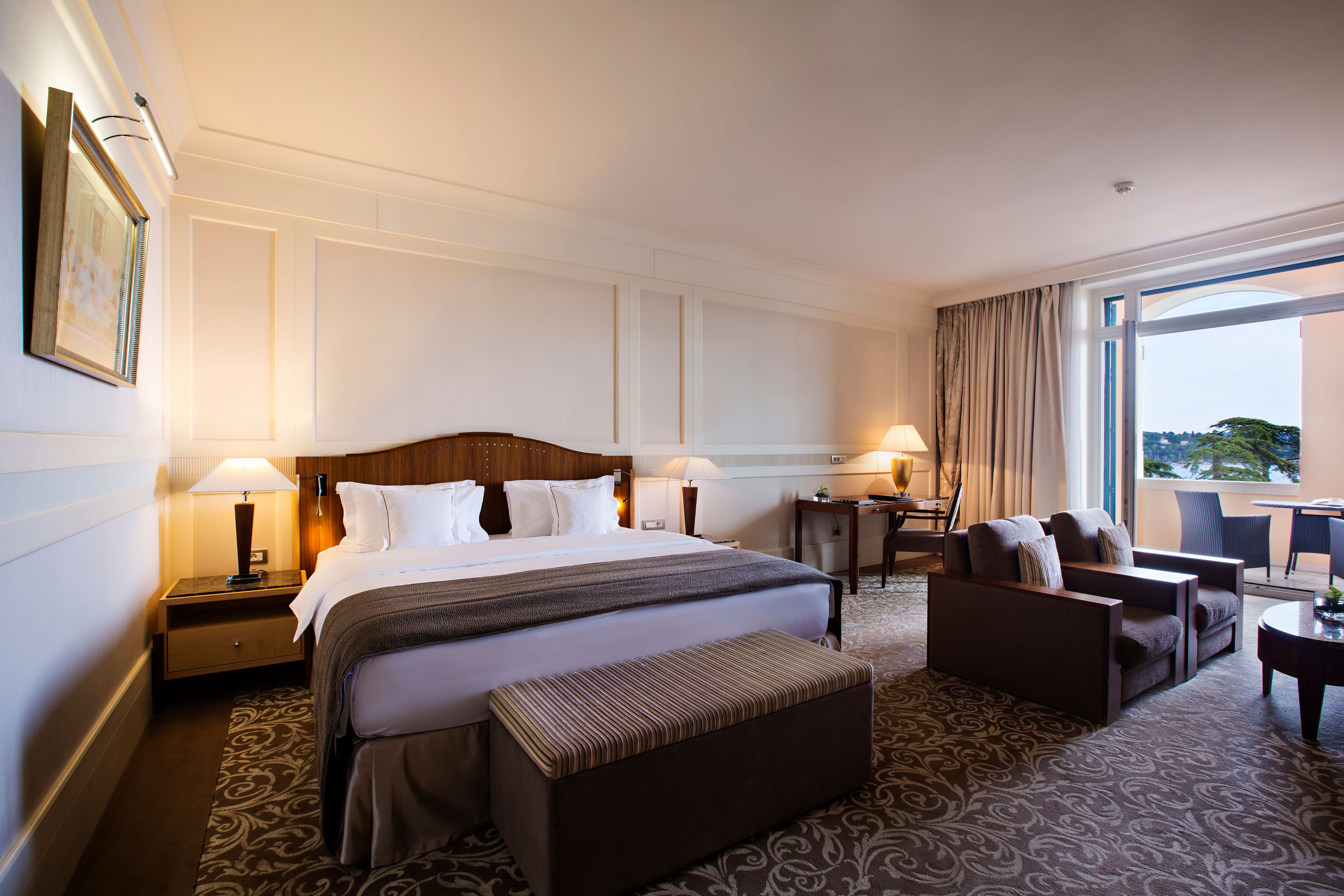 A spacious hotel suite featuring a king bed, a small seating area with two armchairs, a work desk, and open glass doors leading to a balcony with an outdoor view.
