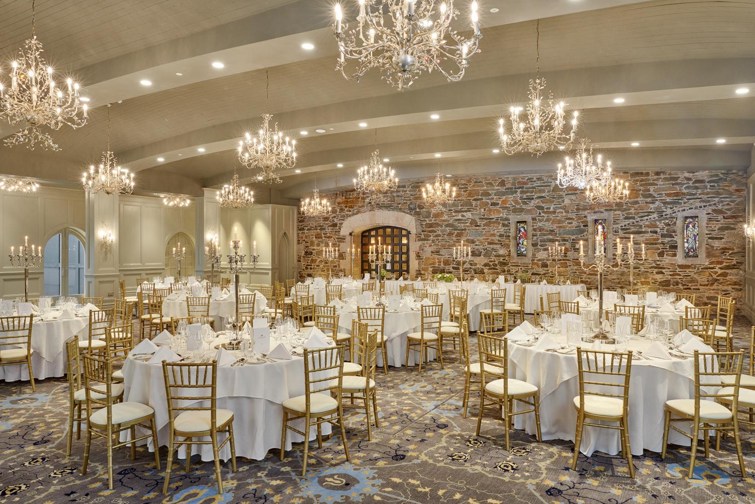 An elegant ballroom set up for a banquet with round tables adorned with white linens and tall candelabras, gold Chiavari chairs, ornate crystal chandeliers, patterned carpet, and an exposed stone wall with decorative archways.