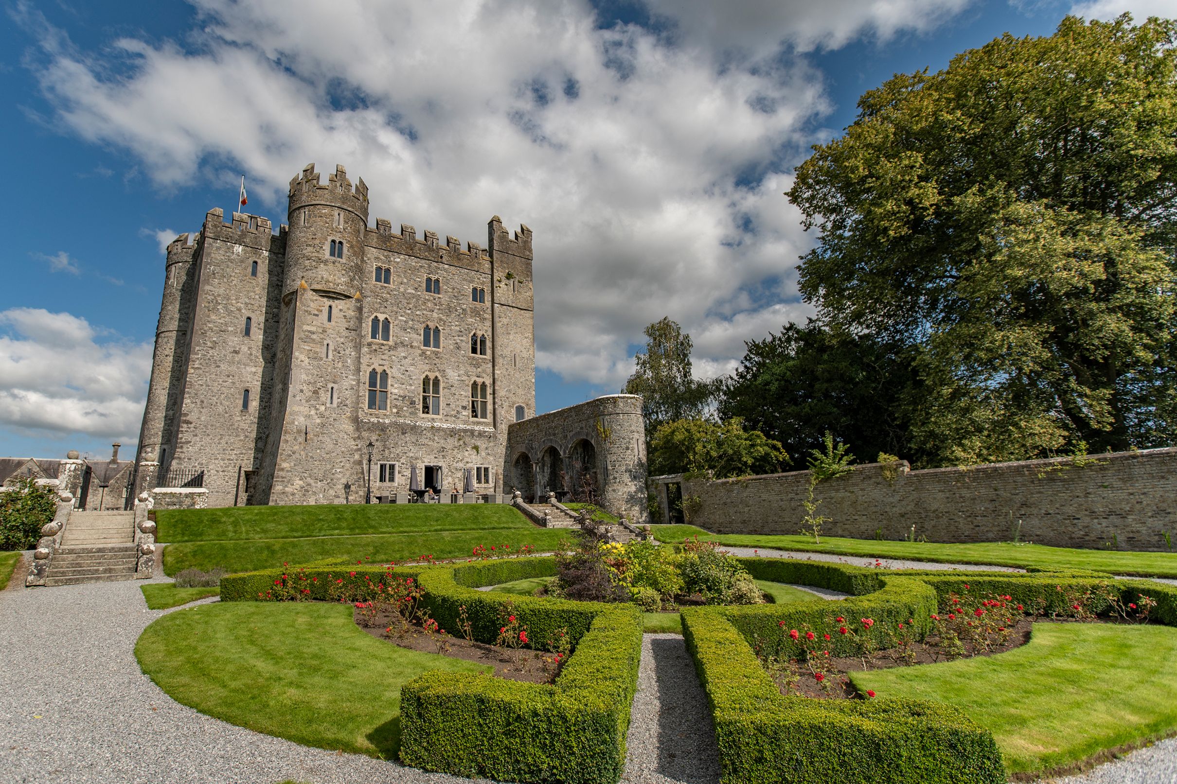A grand grey stone castle with battlemented towers and an Irish flag flying on top, overlooking a formal manicured garden with geometric boxwood hedges, red flowers, and gravel pathways under a partly cloudy blue sky.