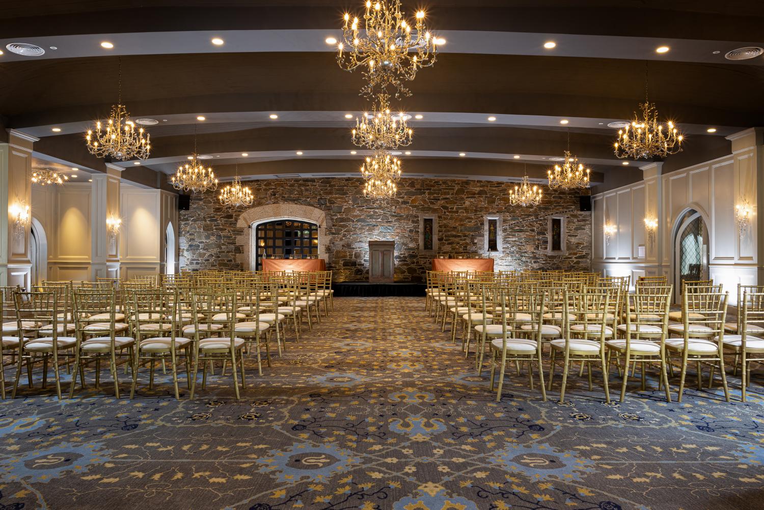 A grand event hall arranged for a ceremony with rows of gold Chiavari chairs facing a podium set against an exposed stone accent wall, lit by crystal chandeliers hanging from a arched dark ceiling.