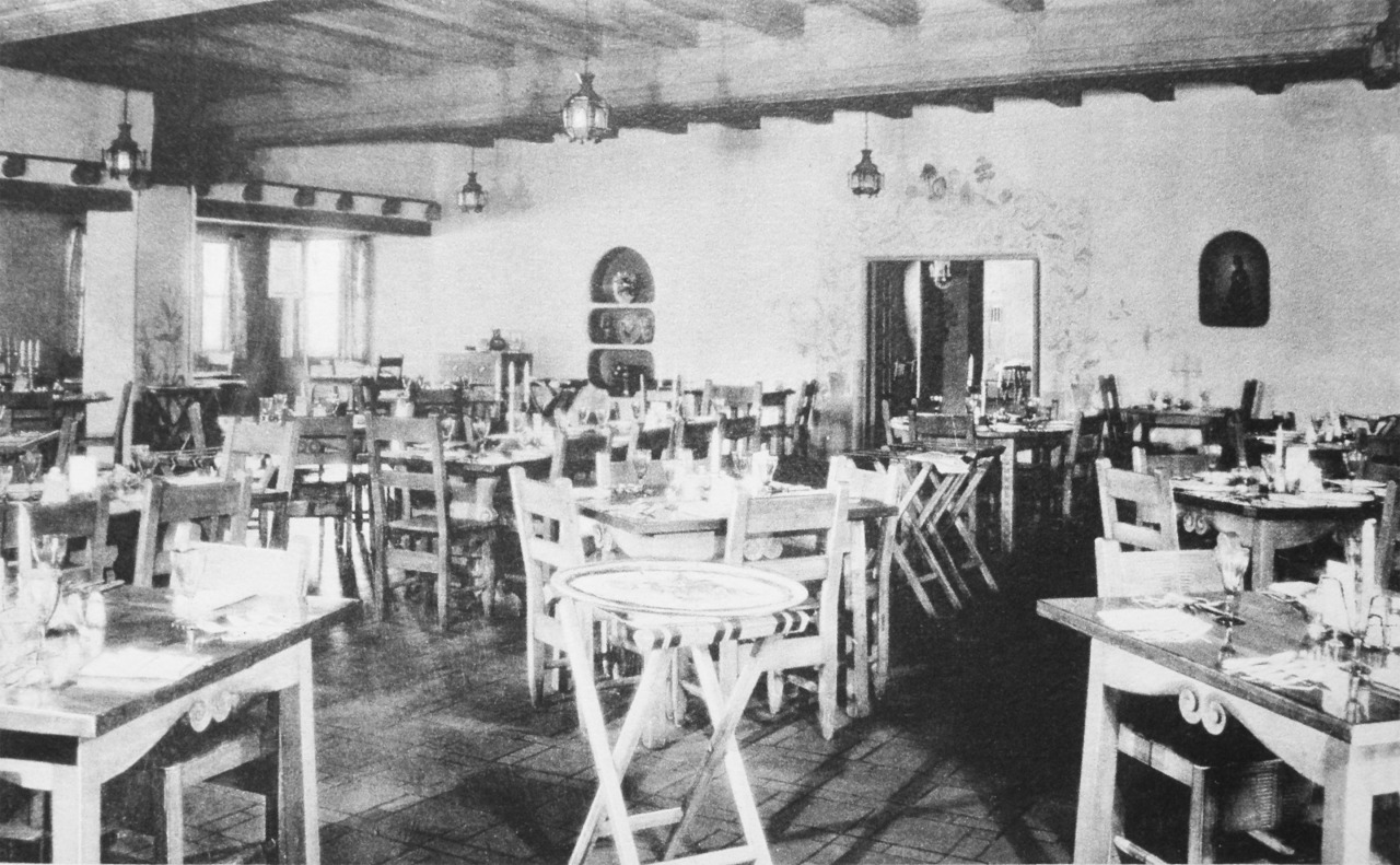 Vintage black-and-white photo of a rustic dining room featuring exposed wooden ceiling beams, hanging lanterns, and simple wooden tables and chairs set for service.