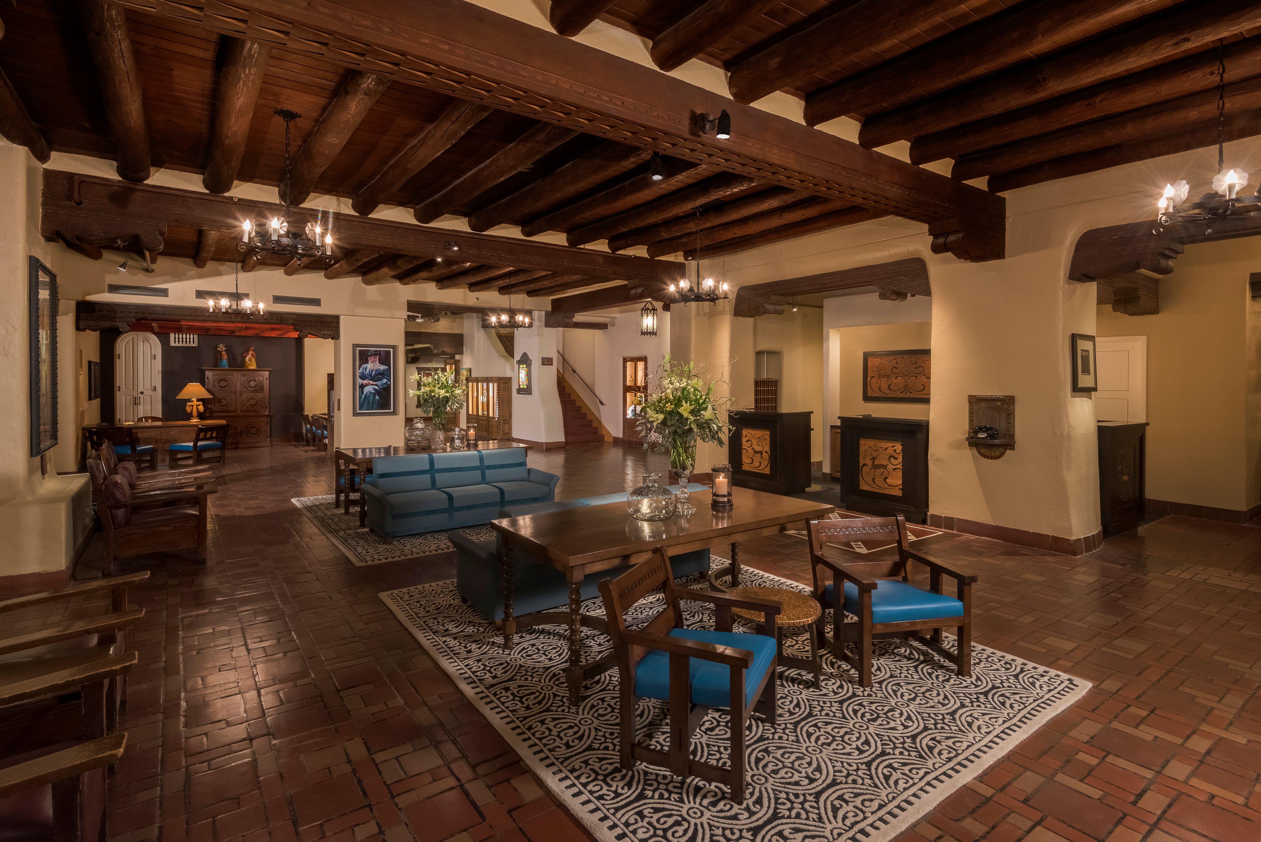 Spacious hotel lobby featuring Southwest Pueblo-style decor with heavy dark log ceiling beams, terracotta tile flooring, patterned area rugs, wooden armchairs, and blue upholstered sofas.