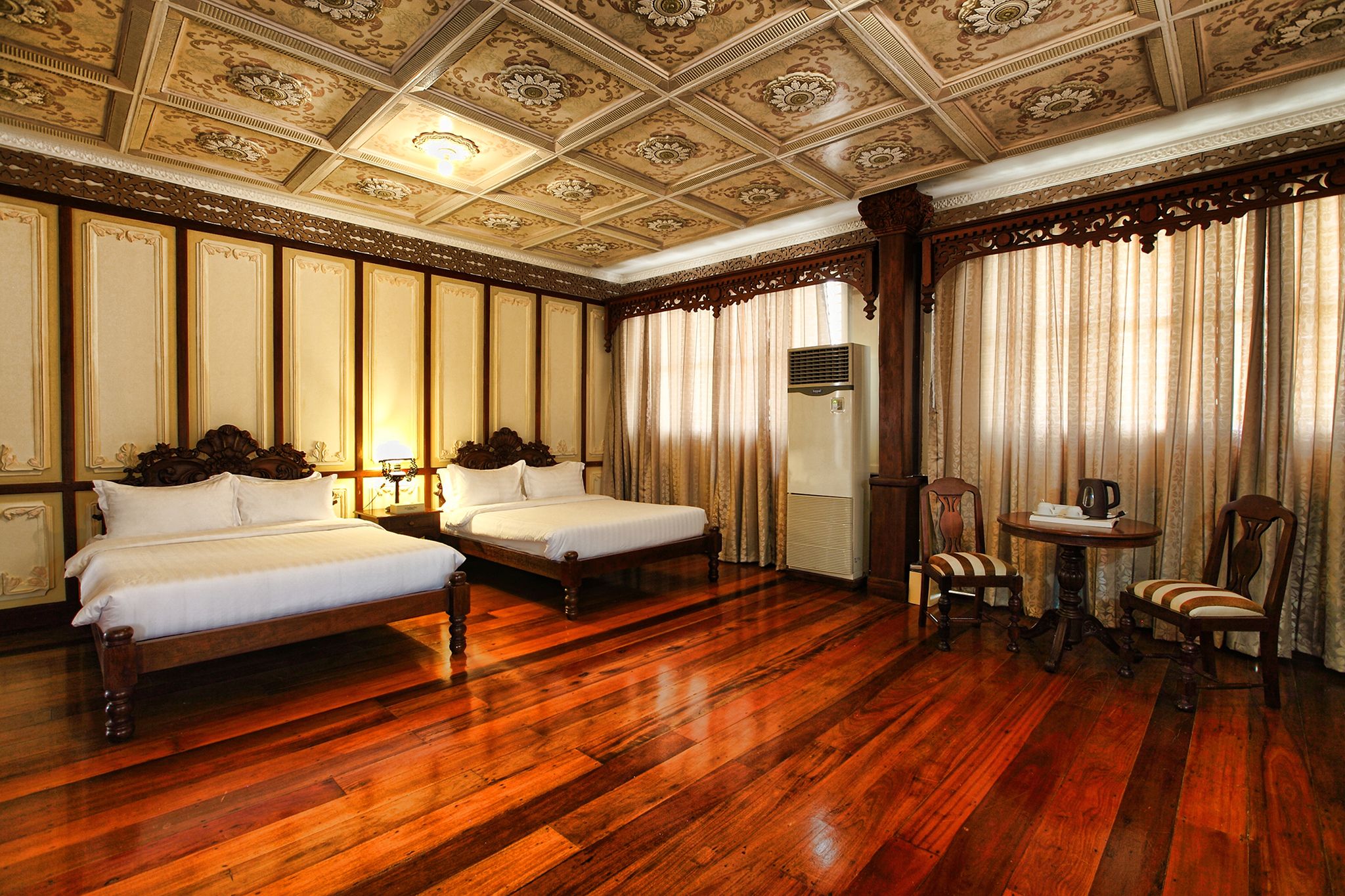 An ornate bedroom featuring two wooden beds with white linens, polished hardwood flooring, intricately detailed ceiling panels with floral medallions, carved dark wood wall trim, and a small seating area with a table and chairs.