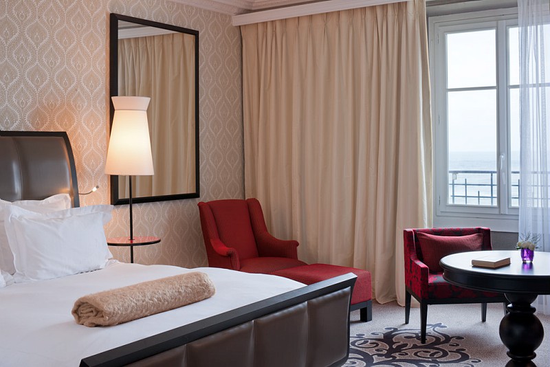 An elegant hotel room interior featuring a bed made with white linens and a rolled beige throw blanket, a red chaise lounge chair, a red patterned armchair beside a round dark wood table, and a large window with cream-colored drapes overlooking the sea.