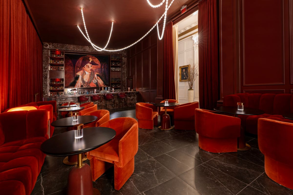 Moody and sophisticated hotel bar with deep red walls, plush orange and red velvet seating, dark marble floors, draped light-string fixtures, and a large artwork displayed behind the bar.