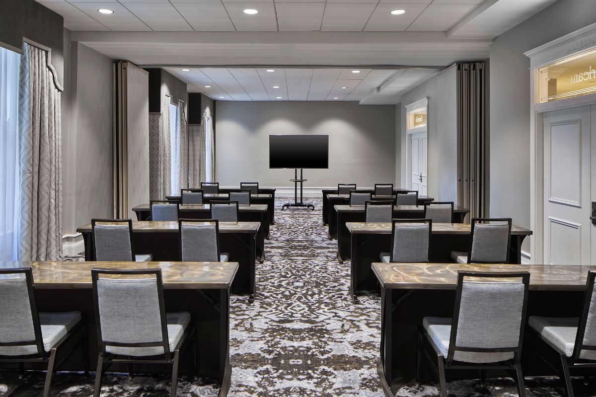 Classroom-style meeting room set with rows of dark wood tables, grey upholstered chairs, a patterned carpet, and a large flat-screen display mounted at the front.