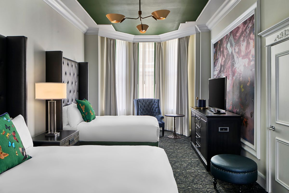 Upscale hotel room with two beds, tall black leather tufted headboards, green butterfly throw pillows, a green recessed ceiling alcove with modern brass lighting, and a bay window sitting area.