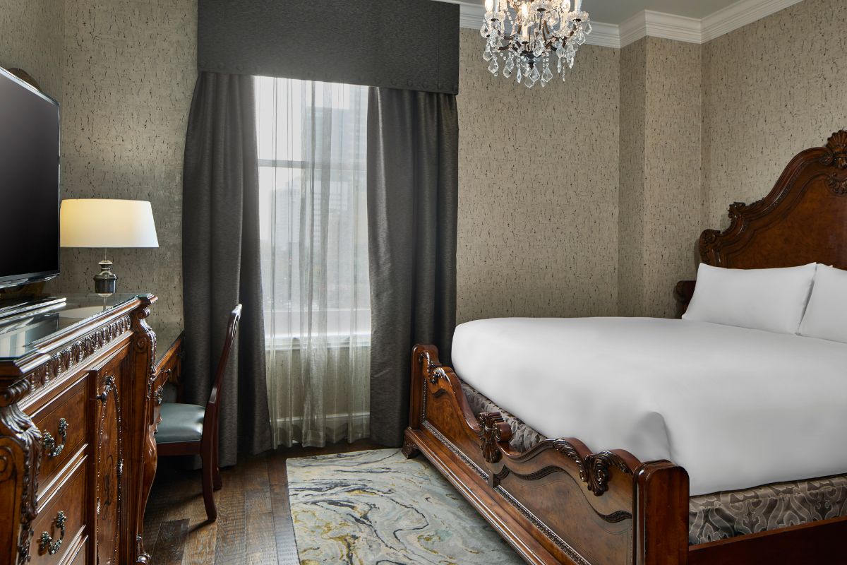 Luxurious guest room featuring an ornate carved wooden bed with crisp white bedding, a matching carved wood dresser with TV, a small crystal chandelier, and dark grey curtains framing a window.