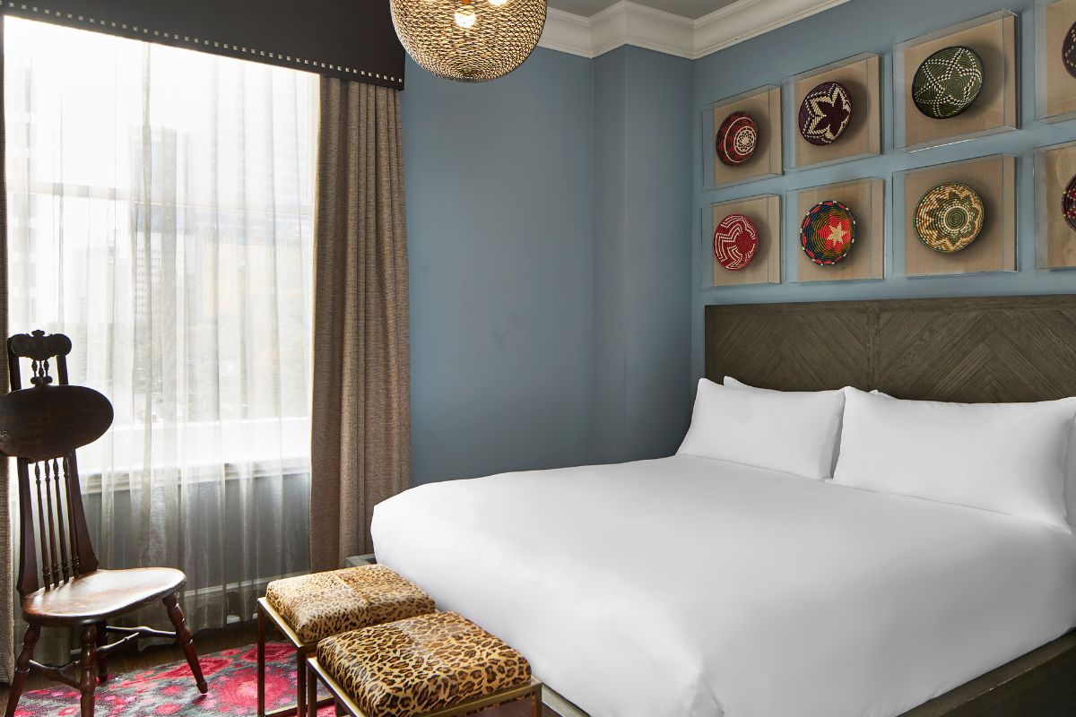 Stylish hotel room with blue walls, a king bed with crisp white bedding, herringbone wood headboard, framed decorative baskets on the wall, leopard-print ottomans at the foot of the bed, and a vintage wooden chair by the window.
