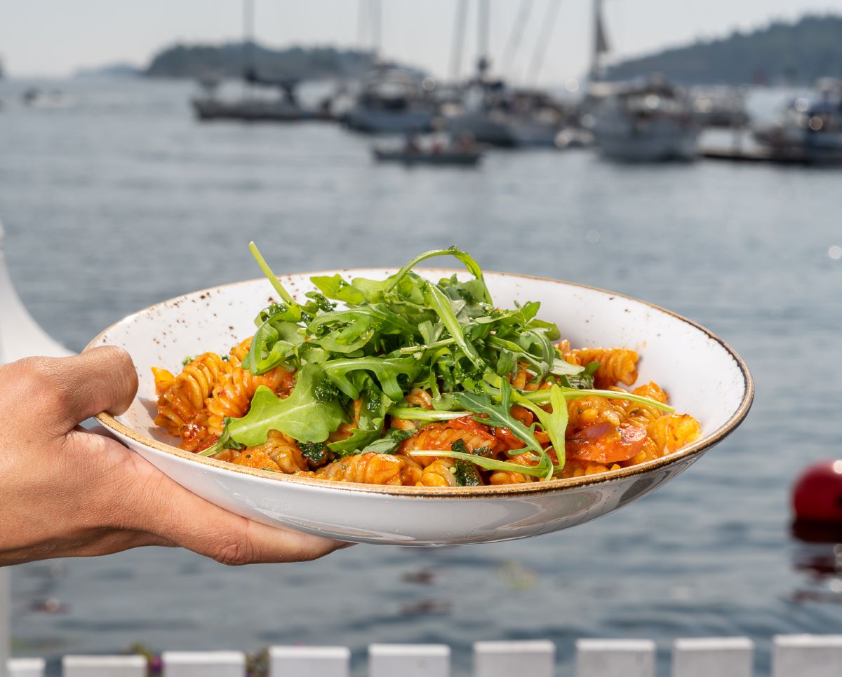 a hand holding a bowl of pasta with a water in the background