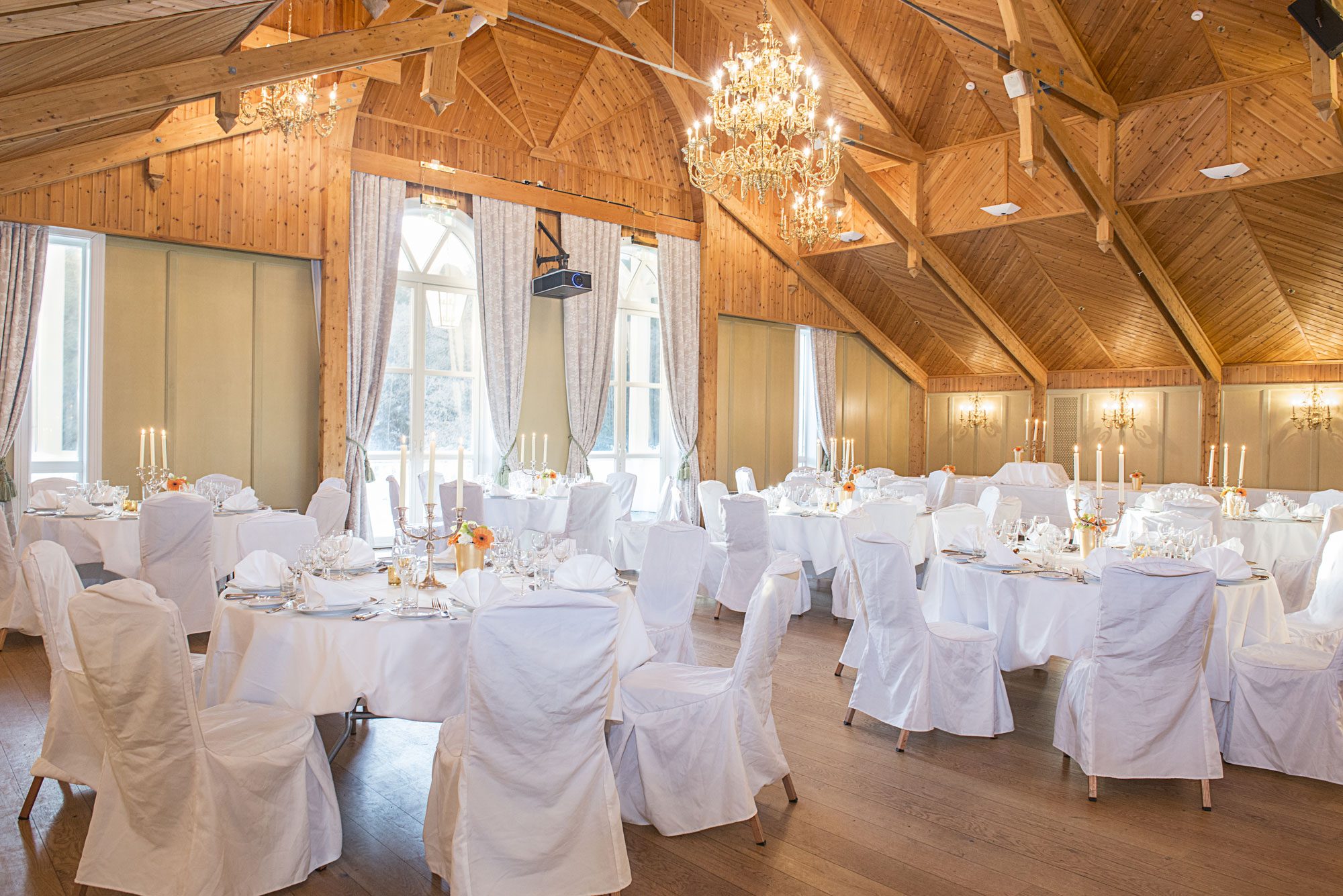 Grand event hall with high vaulted wood-paneled ceilings, exposed timber trusses, and ornate crystal chandeliers, featuring round dining tables set with white tablecloths, white chair covers, tall candelabras, and place settings on hardwood floors.