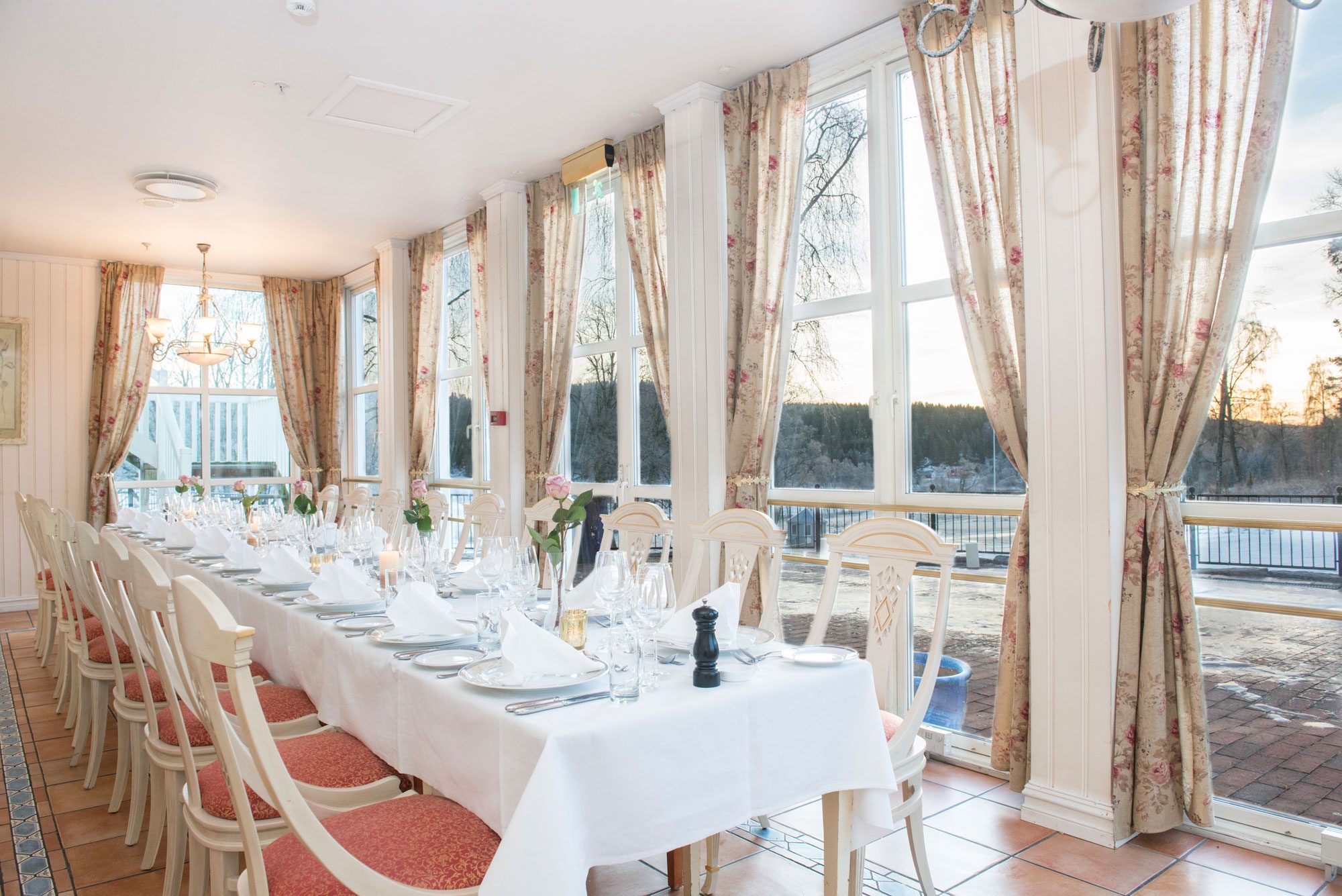 Brightly lit dining room with a long banquet table dressed in white linen, set with fine china and glassware, lined with off-white wooden chairs featuring pink patterned seat cushions, along a wall of large floor-to-ceiling windows with beige floral curta