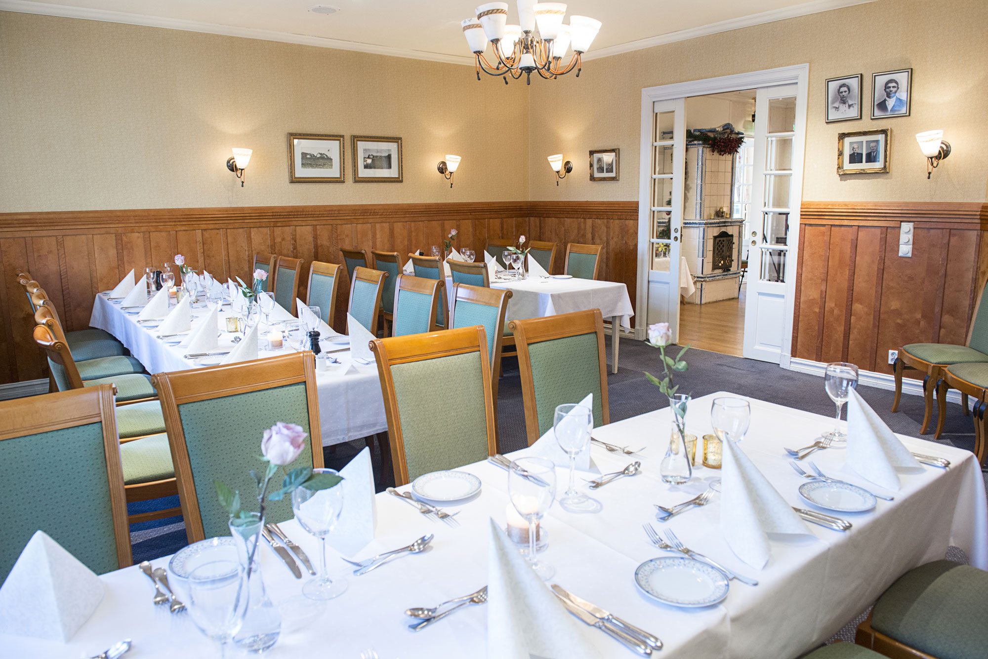 Spacious dining room set up for a event, featuring multiple long tables adorned with white tablecloths, folded napkins, place settings, and pink roses, with green upholstered wooden chairs, wood-paneled walls, wall sconces, and double sliding glass doors 