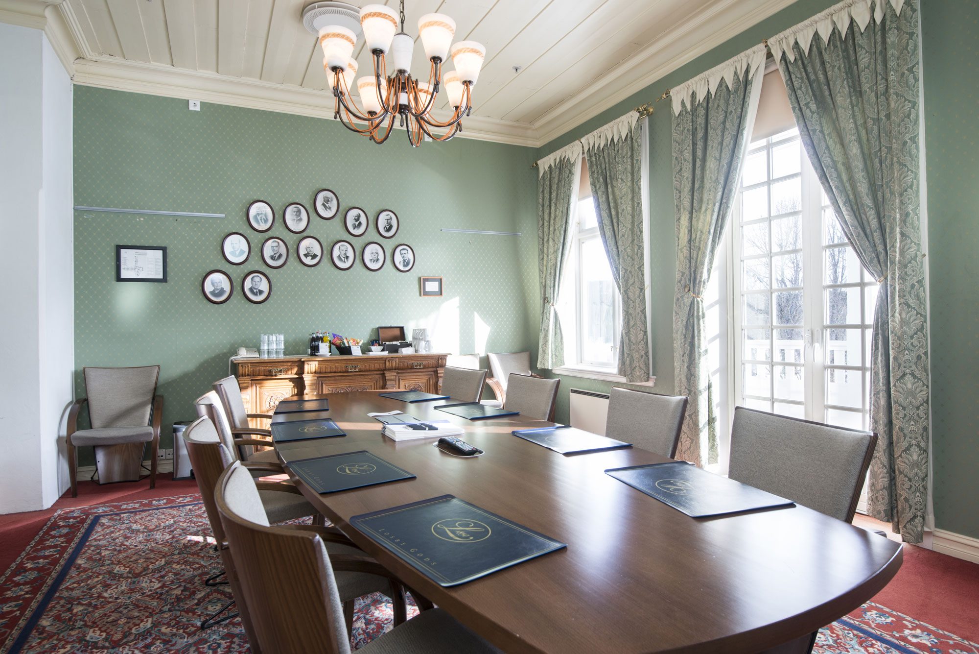 A classic conference boardroom featuring a long oval wooden table set with placemats and notepad folders, surrounded by grey upholstered armchairs on an ornate Persian rug, soft sage-green walls decorated with oval framed portraits, a brass chandelier, an