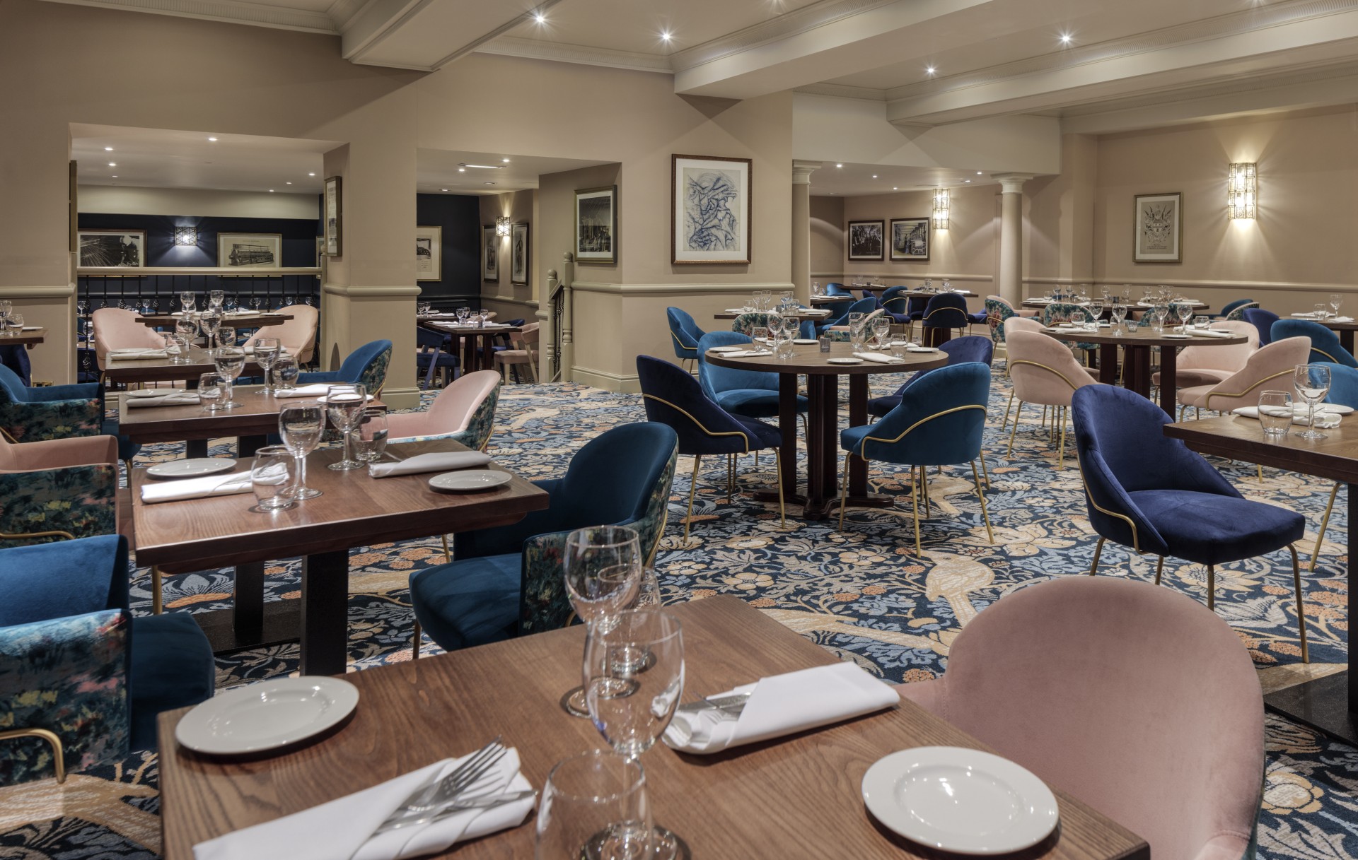 A spacious hotel restaurant featuring wooden dining tables, plush dark blue and soft pink armchairs, intricate patterned carpeting, framed artwork, and warm lighting throughout.