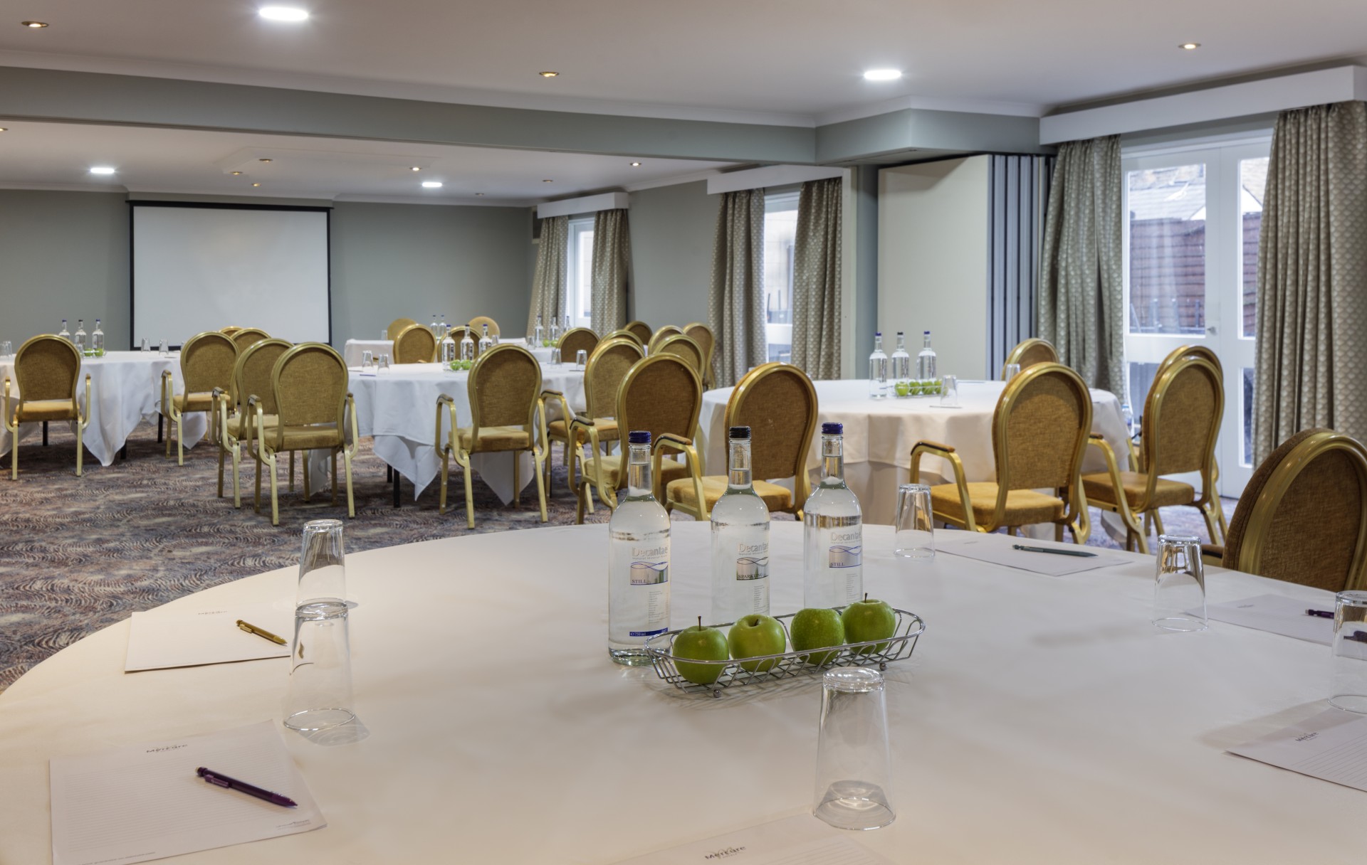 A well-lit meeting room setup with round banquet tables with white tablecloths, gold upholstered armchairs, stationery, bottled water, green apples, and a projector screen at the far end.
