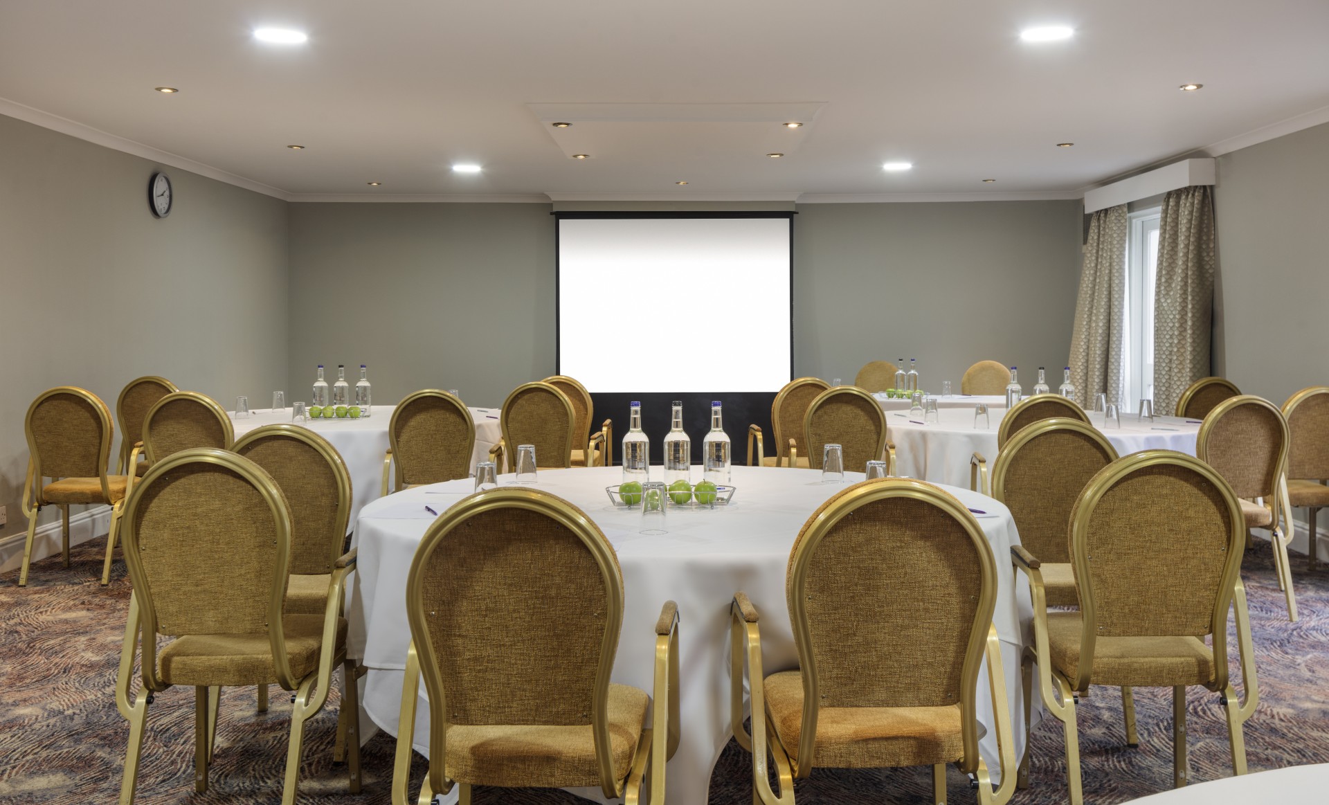 A carpeted meeting room arranged in cabaret style with round tables covered in white tablecloths, gold-framed chairs, glass water bottles, green apples, and a large projection screen at the front.