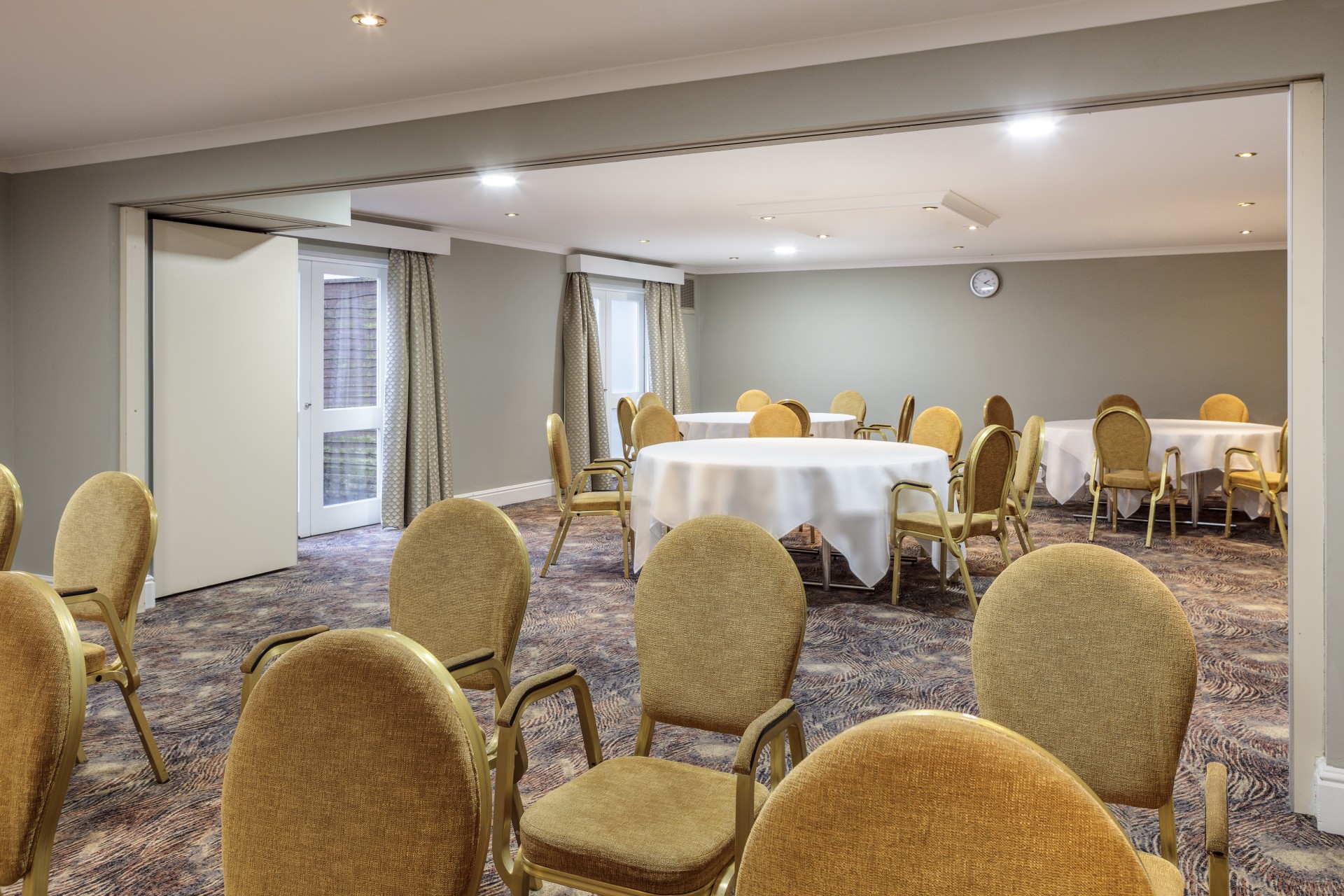 An event space separated by a sliding partition wall, featuring gold upholstered armchairs set up in rows in the foreground and round banquet tables covered in white tablecloths in the background.