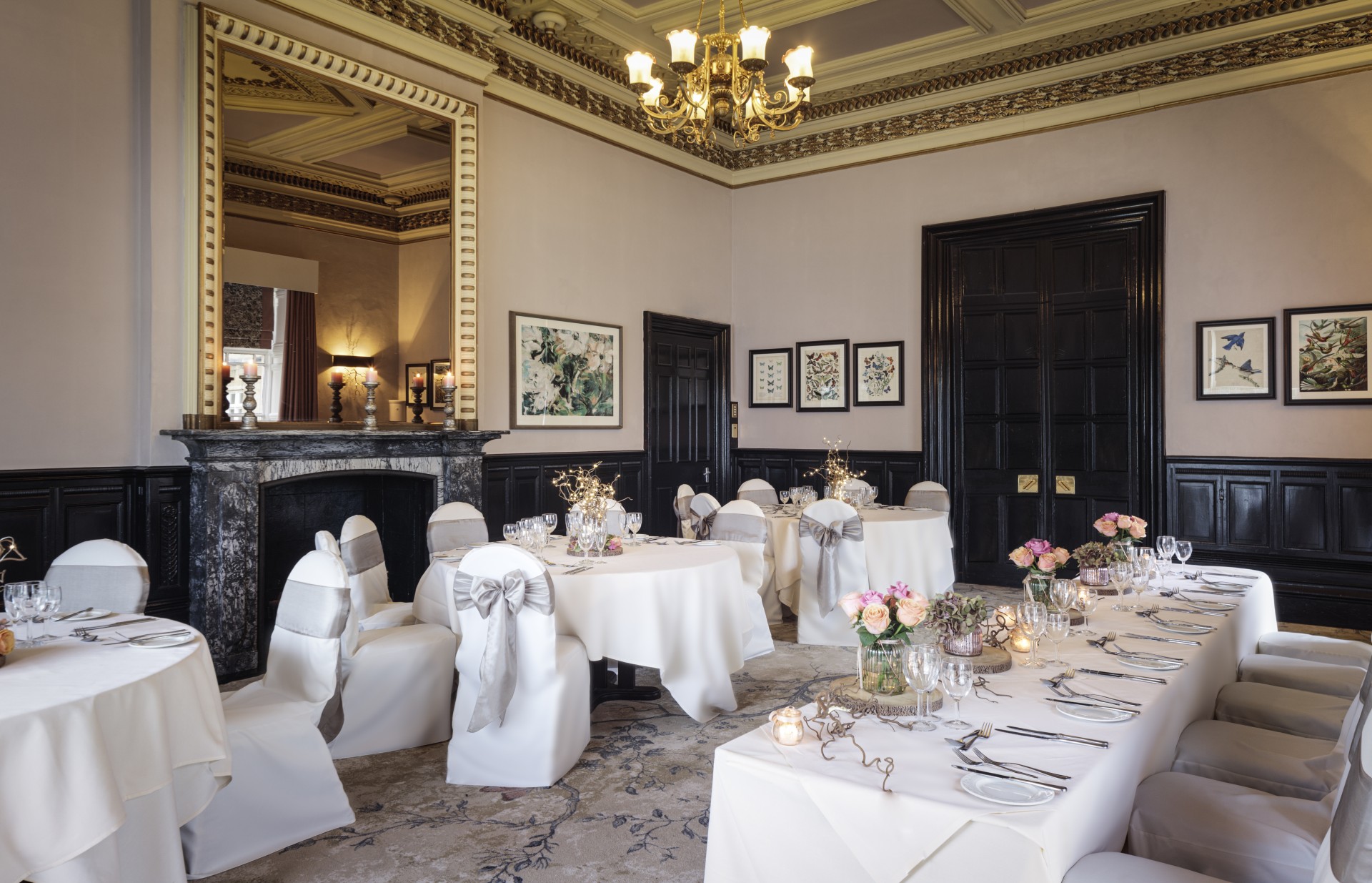 An elegant dining room set up for a special event, featuring a marble fireplace with a large gilded mirror, dark wood panelling, a brass chandelier, and white-lined banquet tables with grey chair sashes and floral centerpieces.