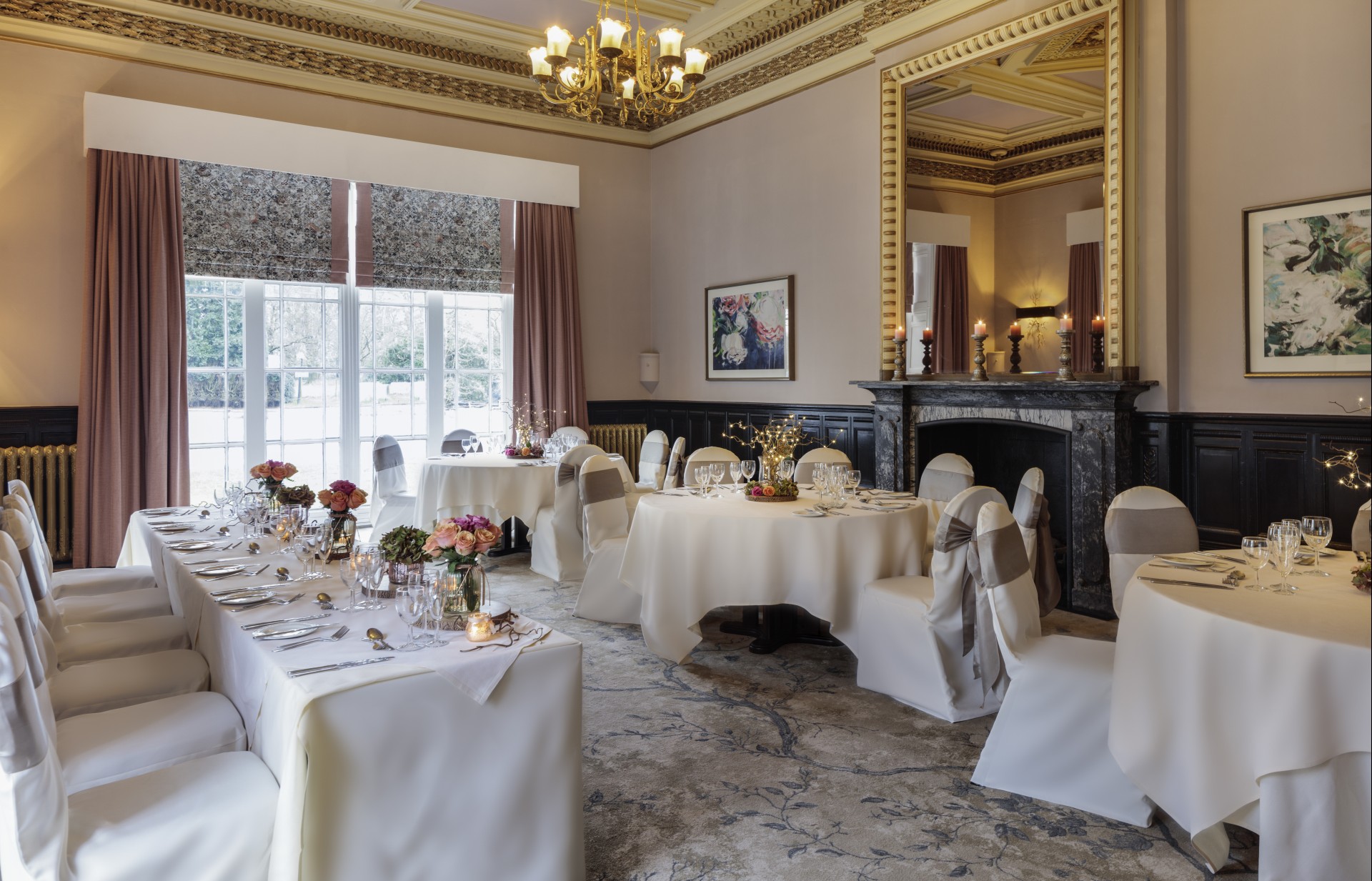 An elegant dining room set up for a special event with white tablecloths, chairs with grey sashes, floral centerpieces, a marble fireplace, and an ornate chandelier.