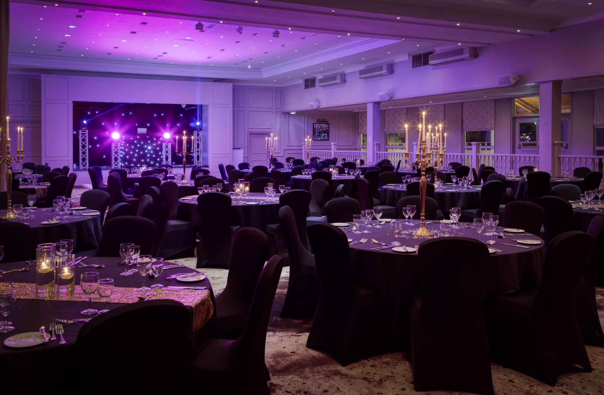 A large event hall set for an evening gala, featuring round banquet tables with black linens and chair covers, gold table runners, candelabra centerpieces, purple ambient lighting, and a stage with lighting rigging in the background.