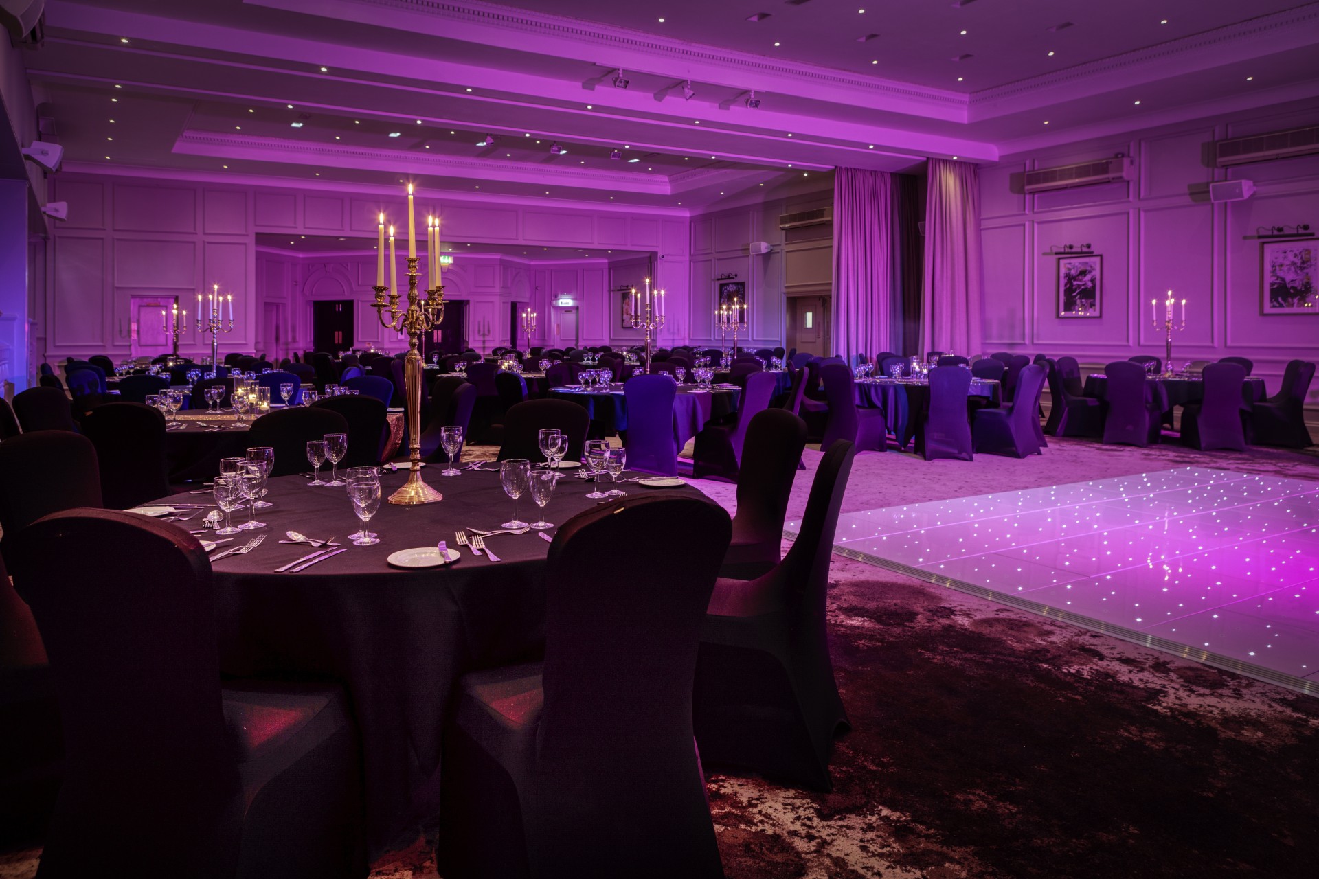 A grand ballroom set for an evening gala or reception, featuring round tables with black tablecloths and chairs, gold candelabra centerpieces, dramatic purple ambient lighting, and an illuminated starlit dance floor.