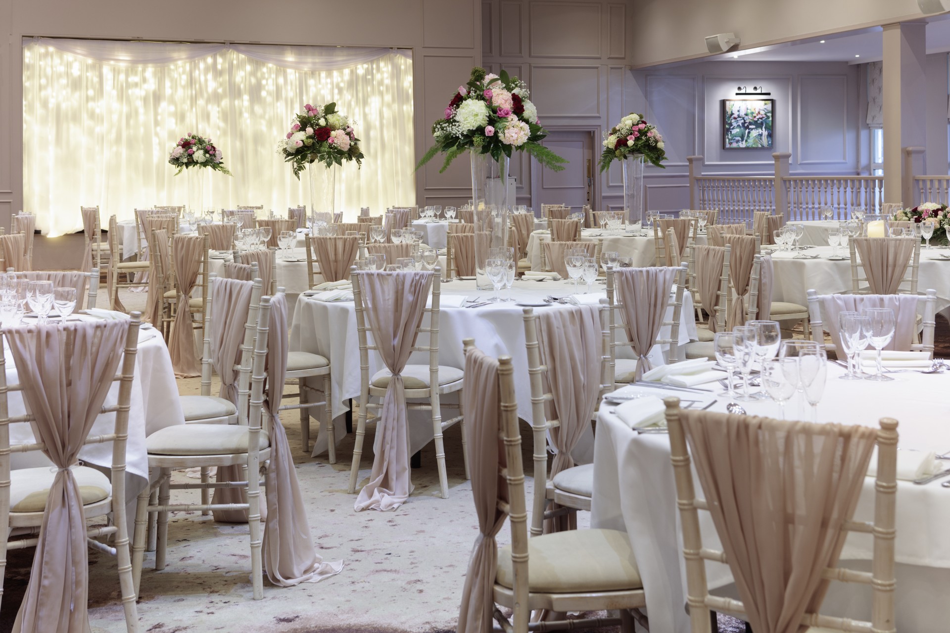A close-up view of round banquet tables in an elegant wedding reception venue, featuring white Chiavari chairs with draped beige sashes, tall floral arrangements, formal place settings, and a fairy light backdrop.