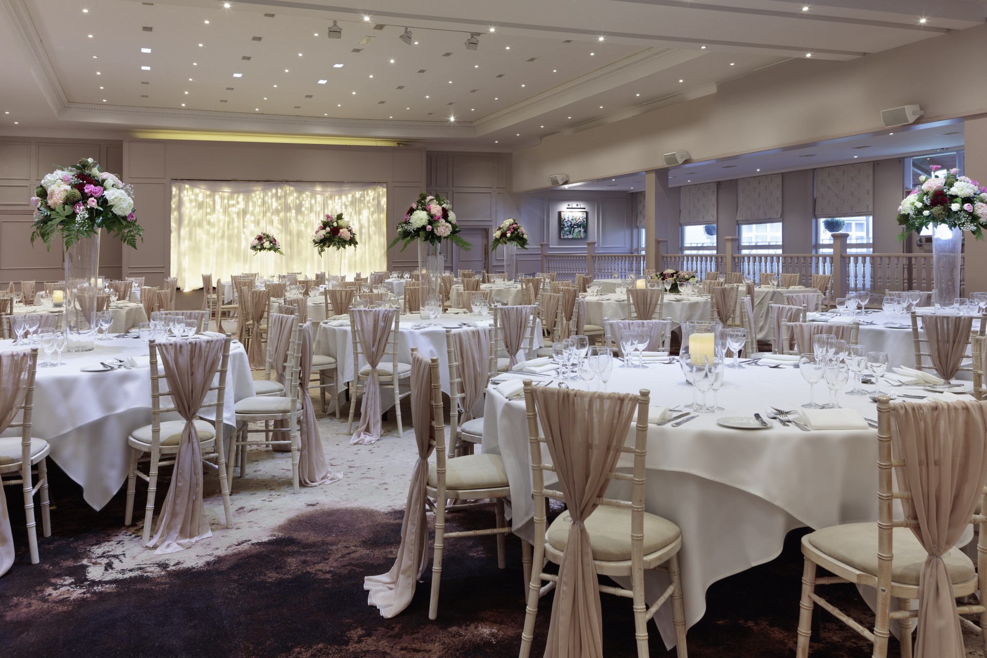 A banquet hall fully set for a wedding reception with round tables covered in crisp white tablecloths, tall floral centerpieces, beige Chiavari chairs tied with matching sheer sashes, and a twinkling fairy light backdrop at the end of the room.