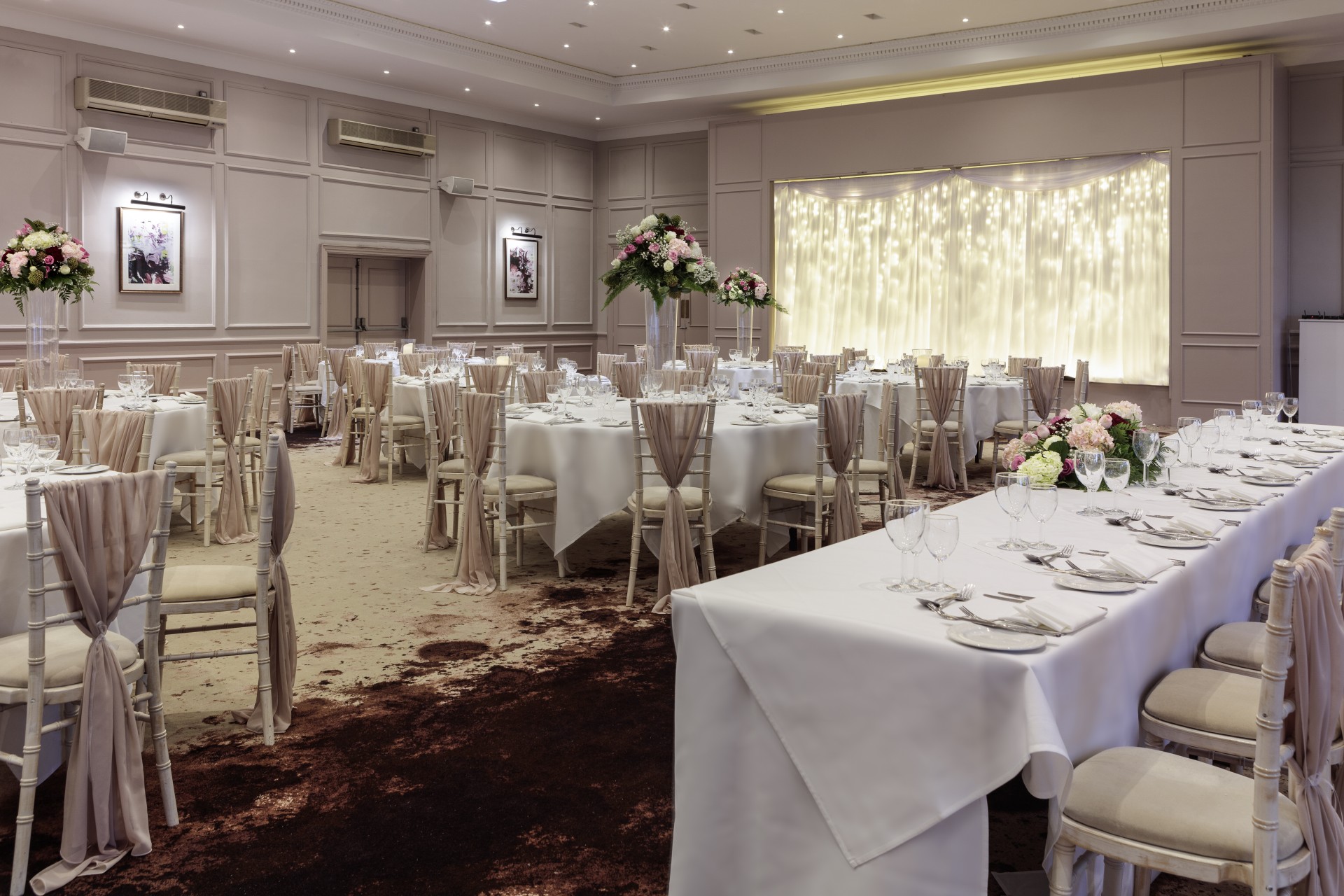 A banquet hall decorated for a wedding reception, with round guest tables, a long head table set with white linens, chairs draped in sheer sashes, floral arrangements, and a illuminated fairy light backdrop on stage.