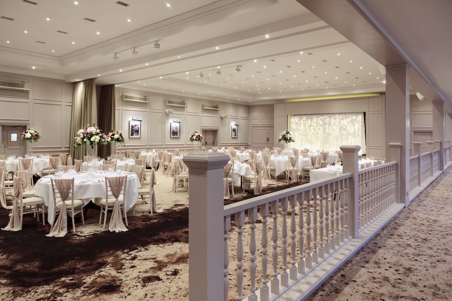 A spacious, brightly lit event hall arranged for a wedding reception, featuring round tables with white linens, floral centerpieces, chairs with neutral sashes, and a fairy light backdrop on a distant stage.
