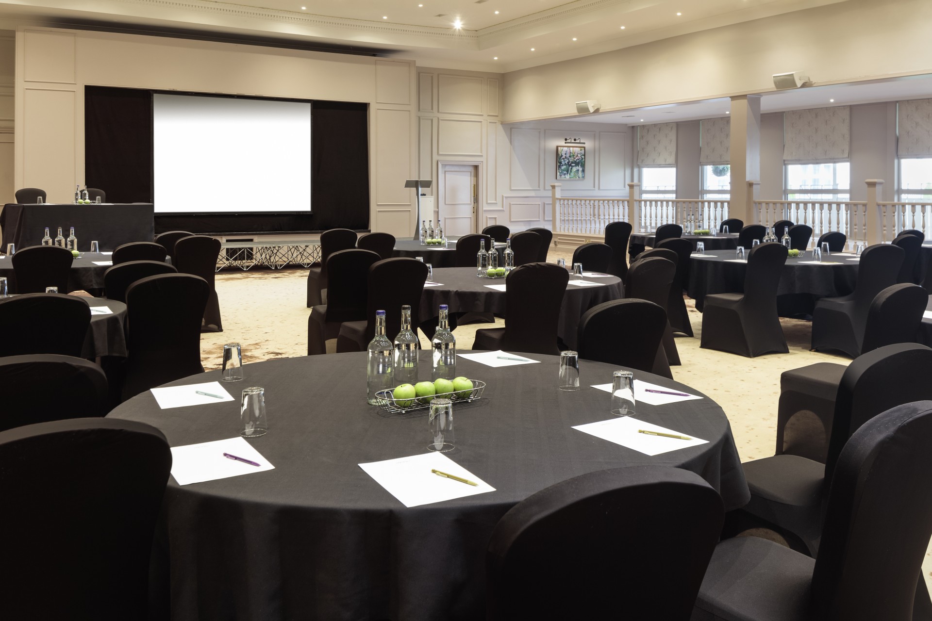 A large conference room set up cabaret-style with round tables covered in black linens, black chair covers, notepad place settings, water bottles, green apples, and a large projection screen at the front.