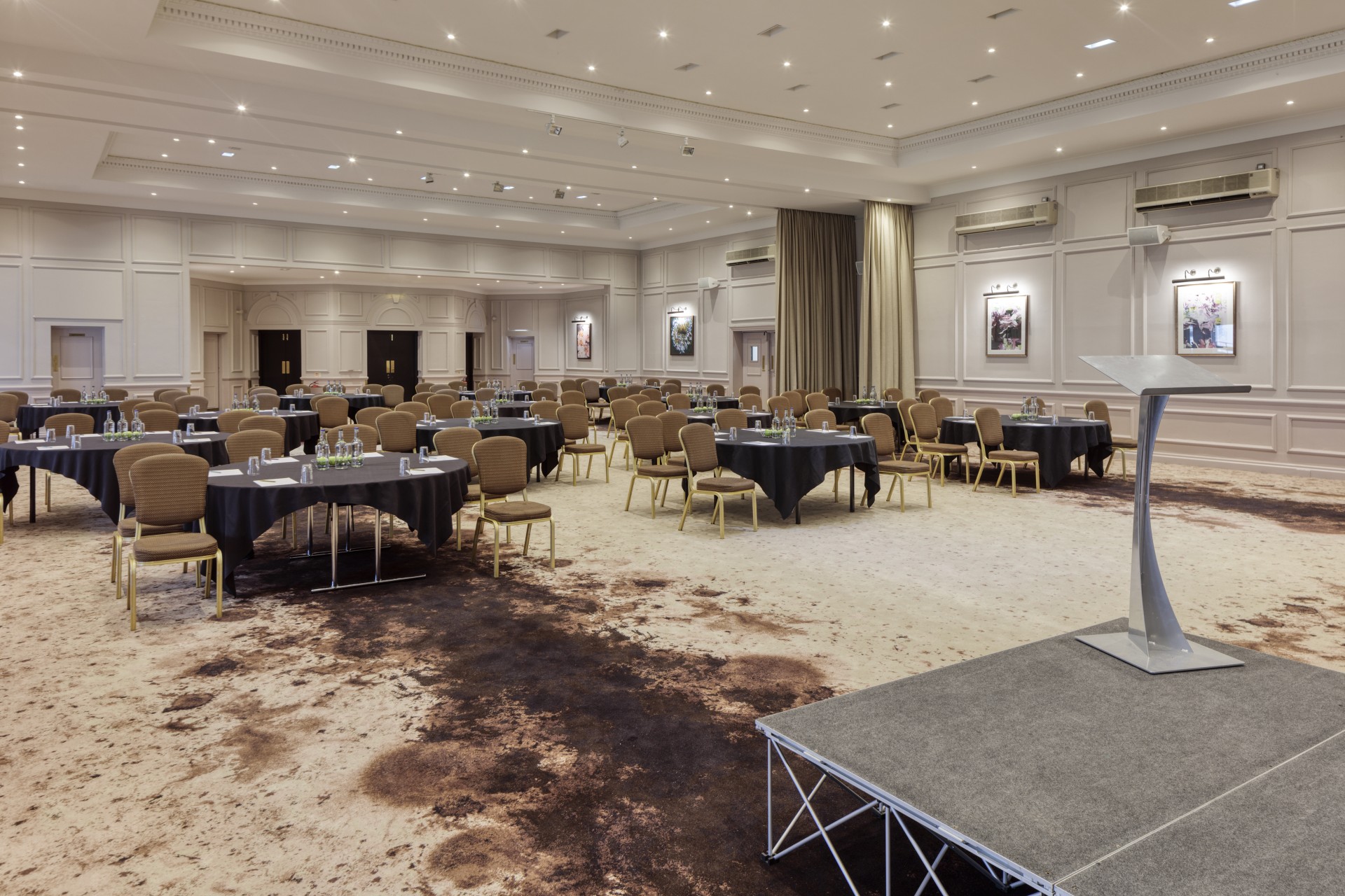 A grand ballroom set up cabaret-style with round tables draped in black tablecloths, gold upholstered chairs, a small carpeted stage with a podium in the foreground, and recessed ceiling lighting.